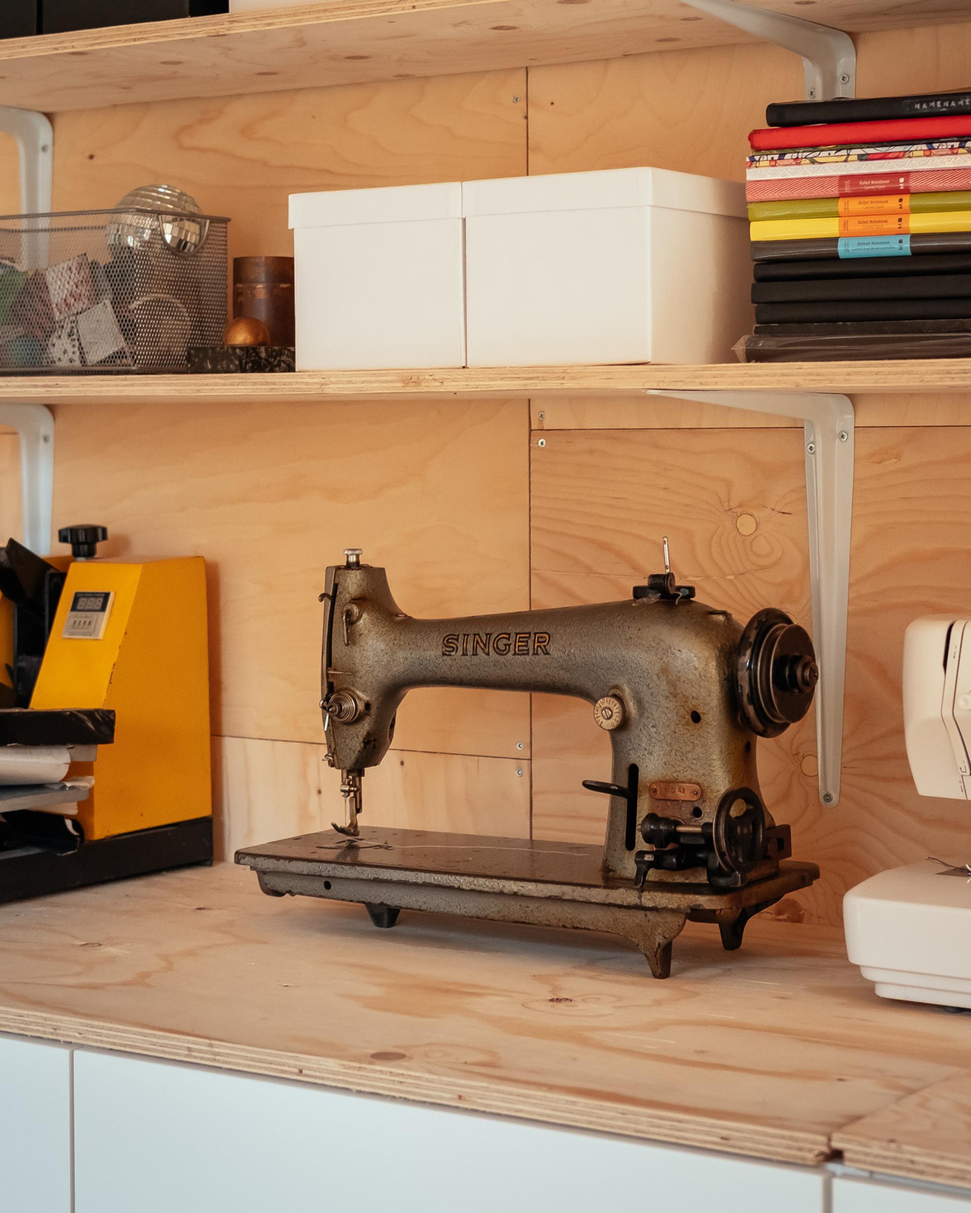 An old Singer sewing machine sits on a wooden shelf beside a yellow device, white boxes, and stacked books, inviting inspiration to upcycle in a light wood space with wall-mounted shelving.