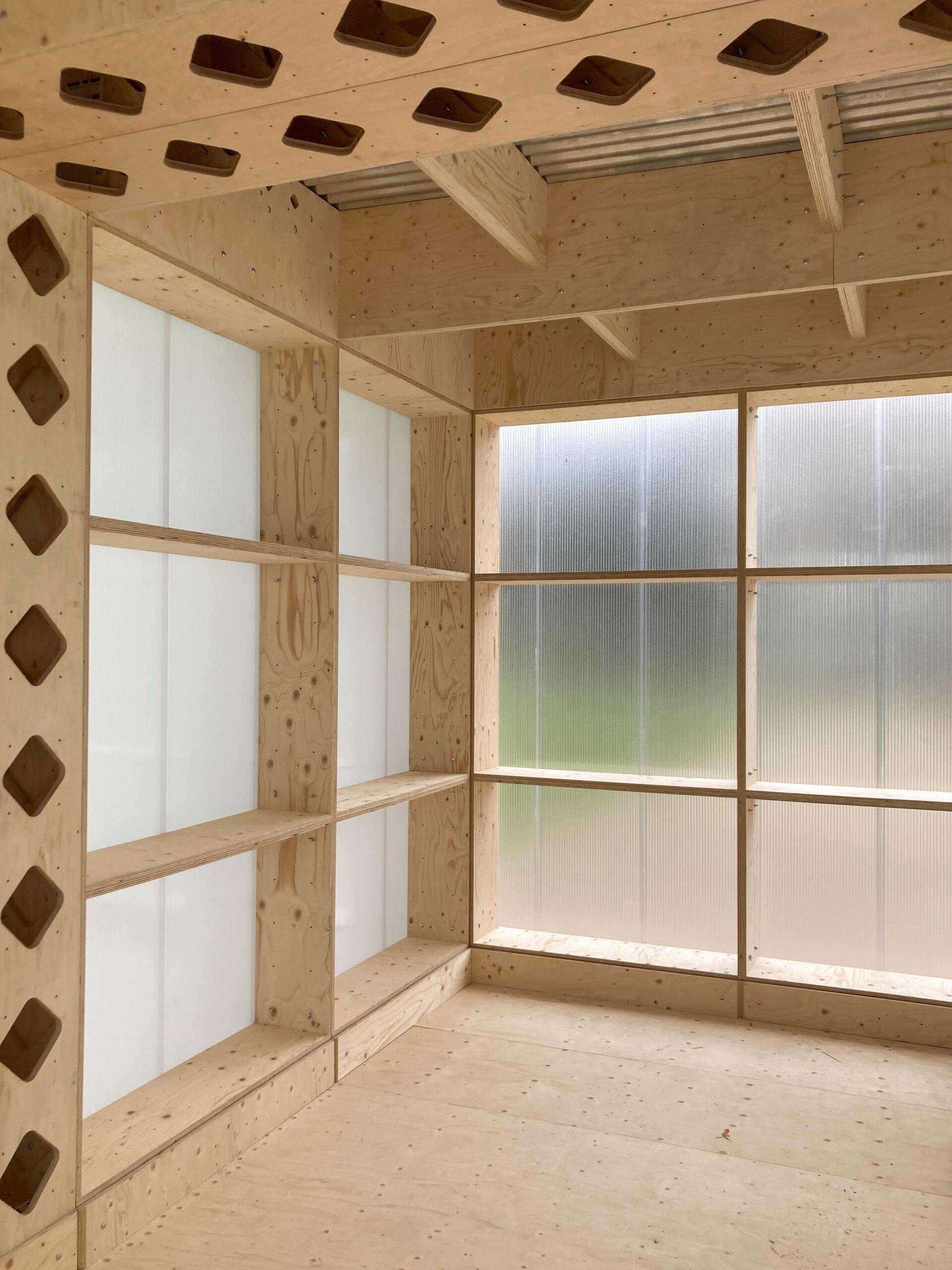 A room with unfinished spruce walls, built-in shelves, and large frosted windows letting in diffused light. The ceiling has exposed beams and geometric cut-outs on the left side.