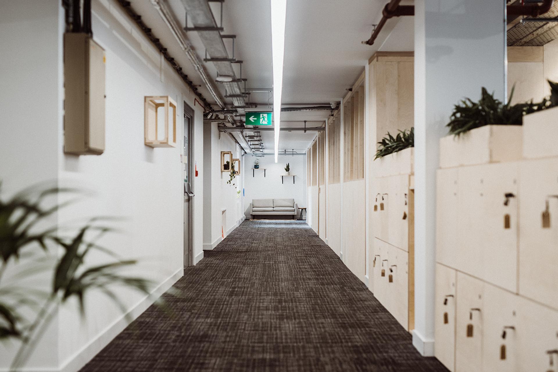 A modern office corridor features sustainable timber construction with white walls, grey carpet, wooden lockers on the right, and potted plants. A beige sofa sits beneath an illuminated green exit sign at the corridor’s end.
