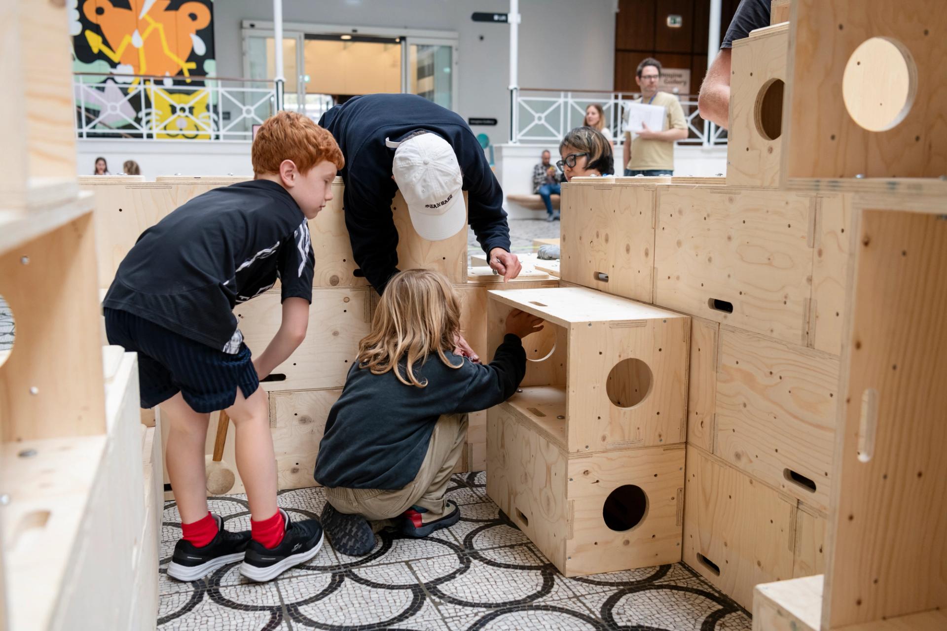 Three children interact with large U-Build wooden building blocks, assembling and exploring them in a public indoor space. Other people and abstract art are visible in the background.