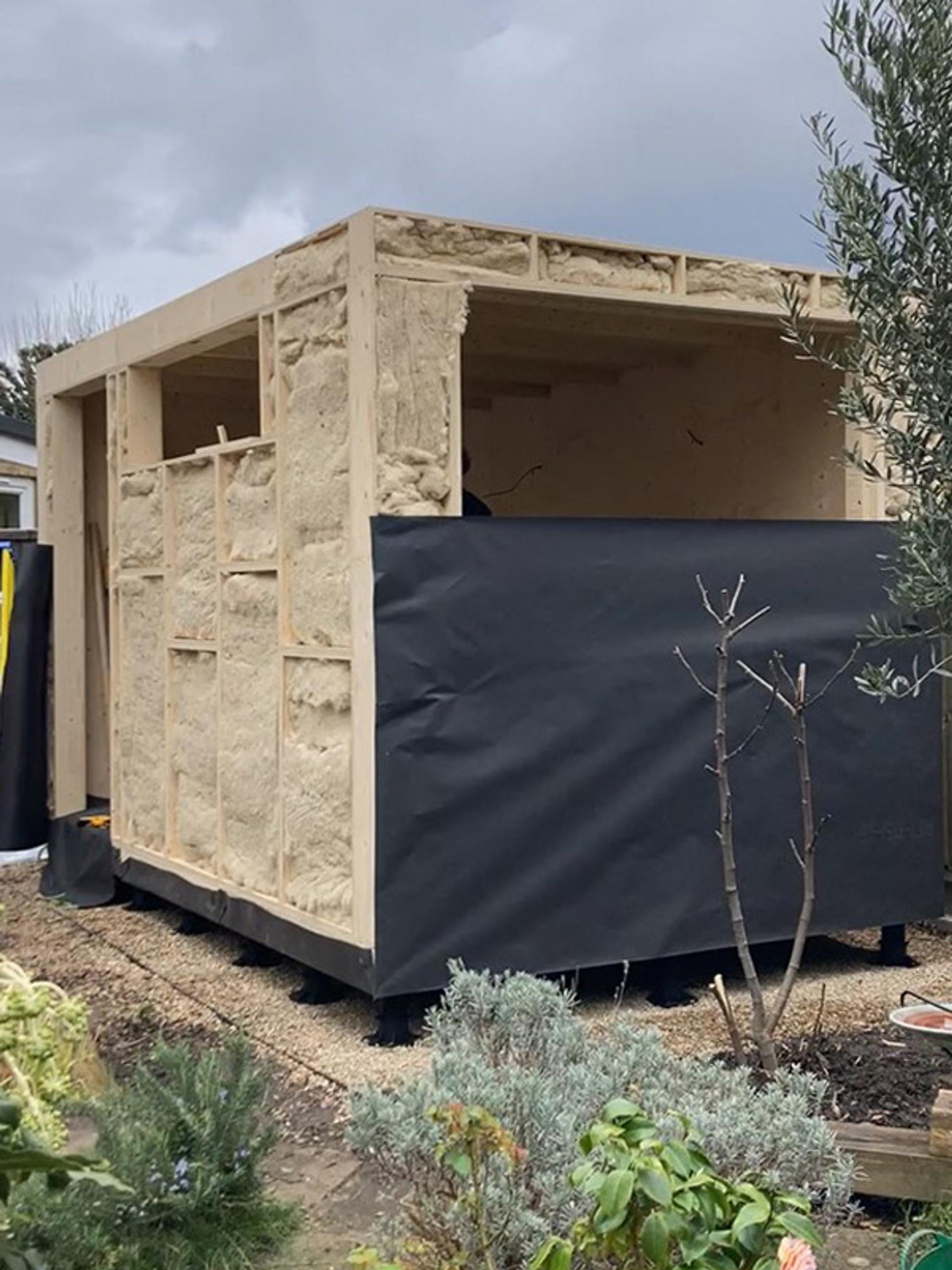 A small timber construction with exposed insulation and partially covered in black weatherproof sheeting stands in a garden, its plywood structure visible amid plants and shrubs in the foreground.