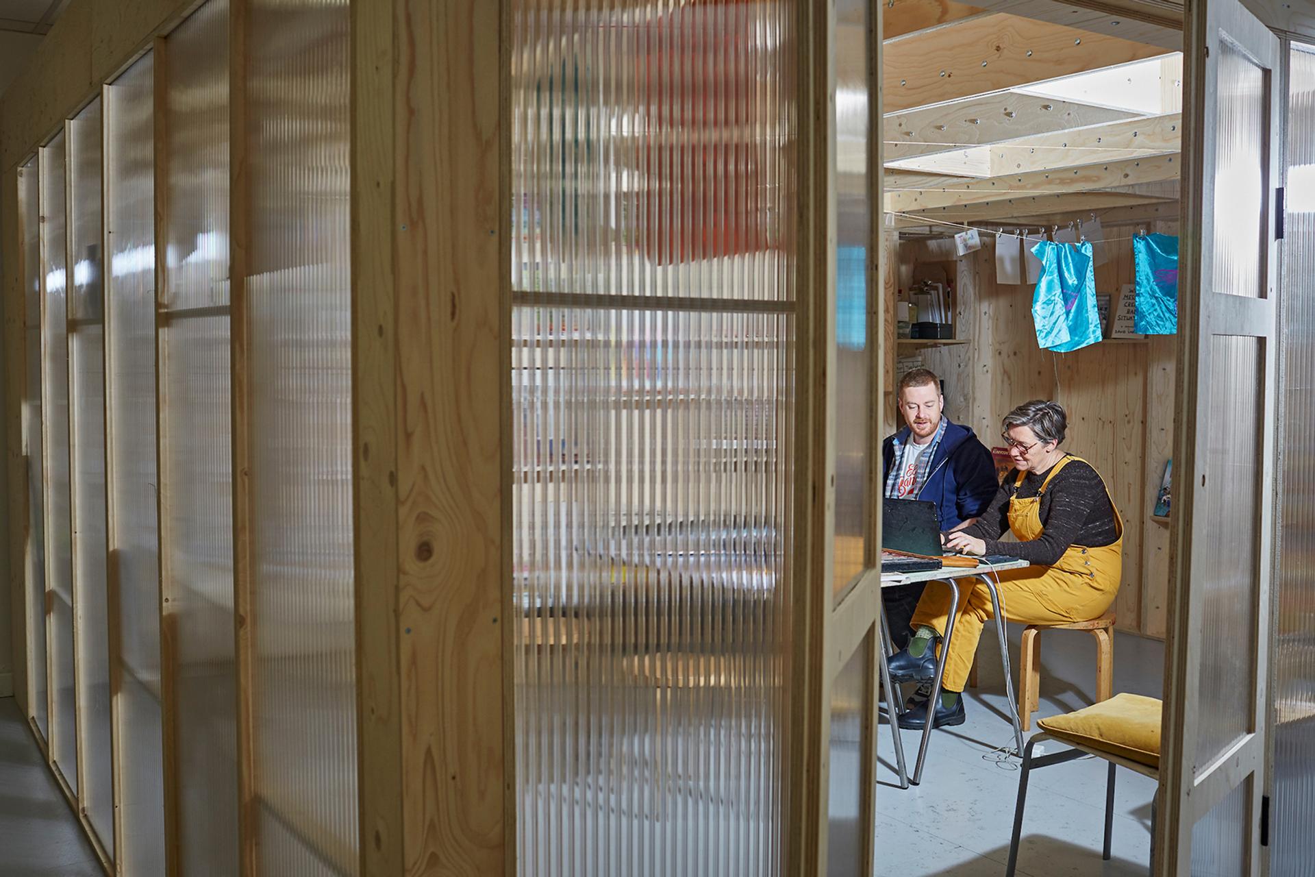 Two people sit at a small table in a modern room with modular birch plywood walls, working together on a laptop and papers. Bright clothing and colourful items hang in the background.