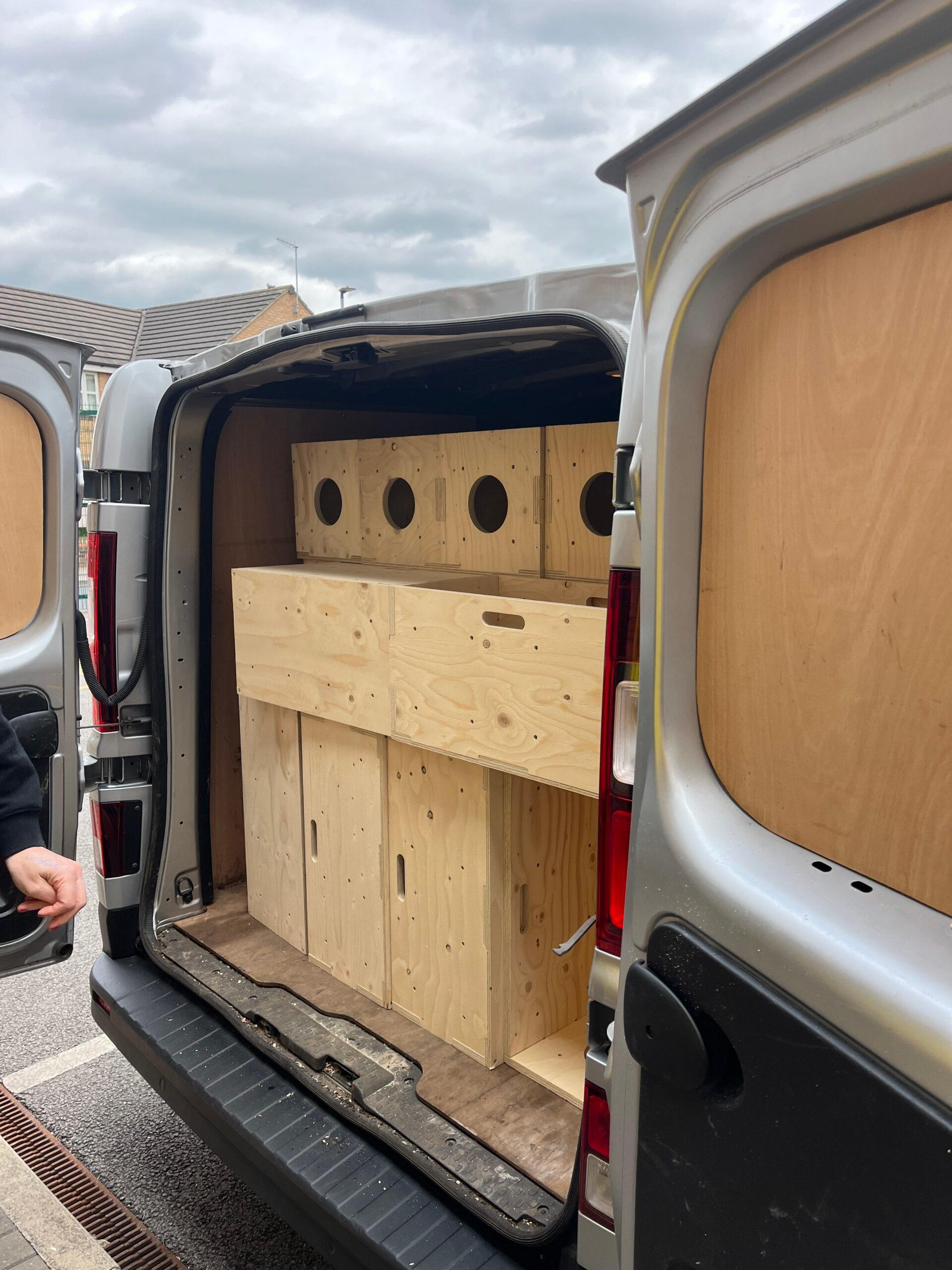 The open back of a van revealing several large spruce wooden boxes with circular holes and handles, neatly stacked inside. A person's arm is visible at the left edge of the image.
