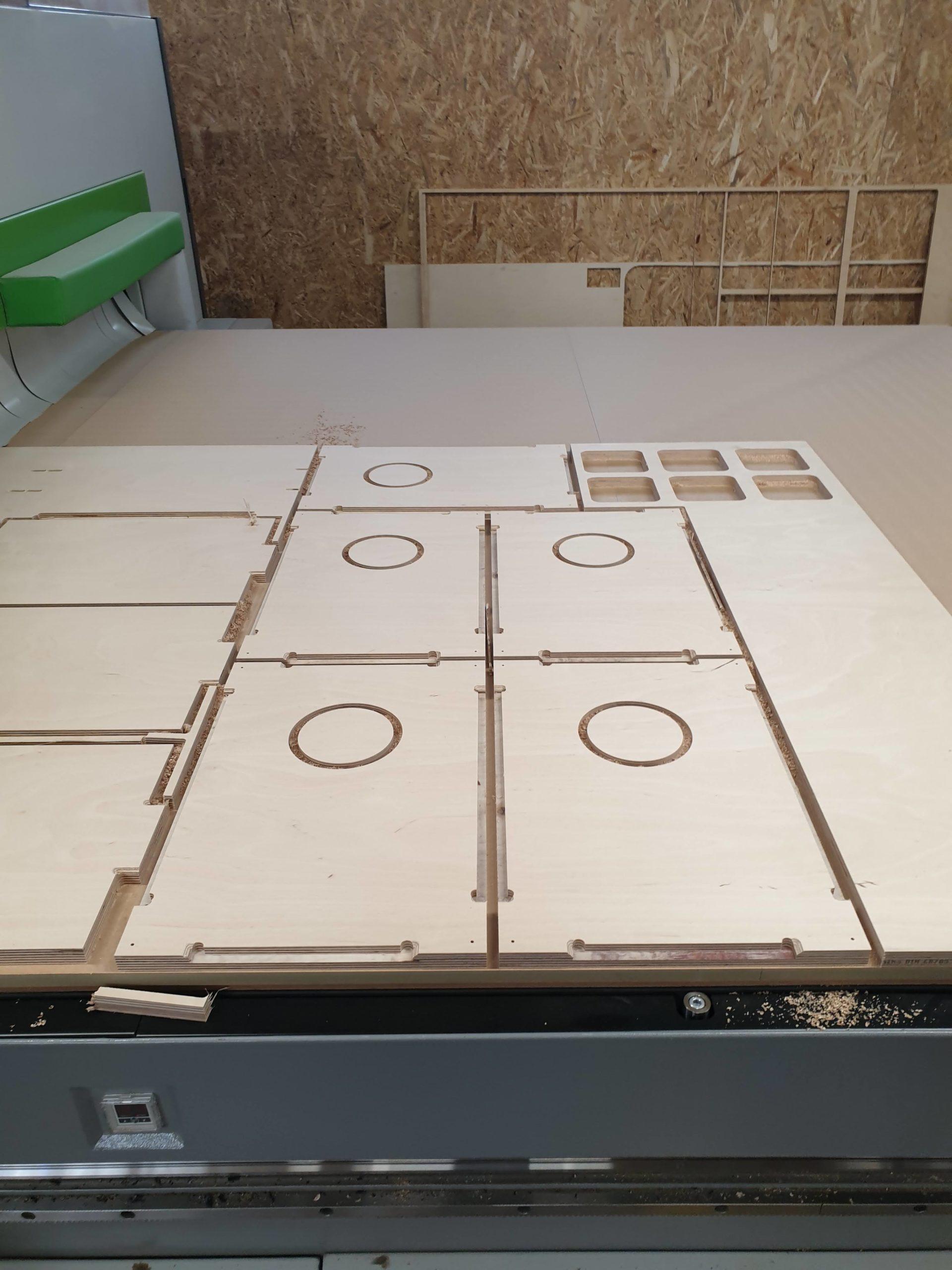 A CNC machine is precisely cutting multiple shapes into large birch plywood sheets in a workshop, surrounded by wooden walls and panels.