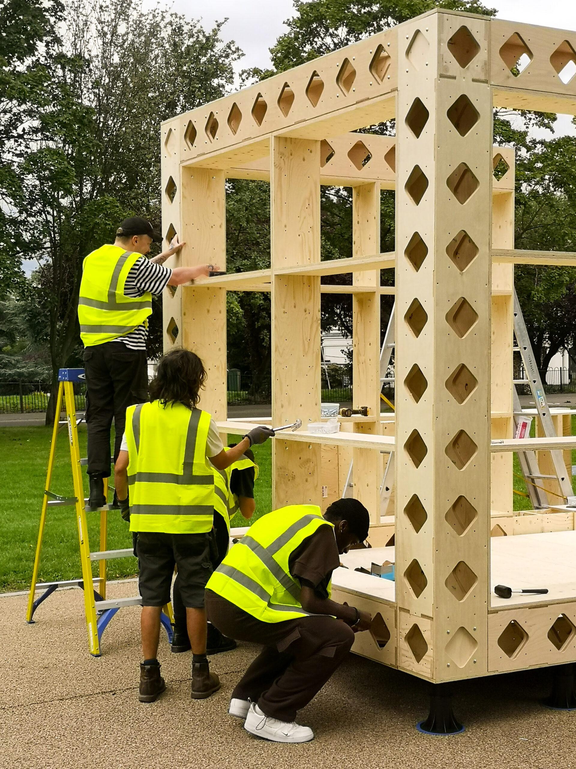 Four people in yellow safety vests work together to assemble a large modular wooden structure outdoors. One person stands on a ladder, while others use tools at ground level. Trees and grass are visible in the background.