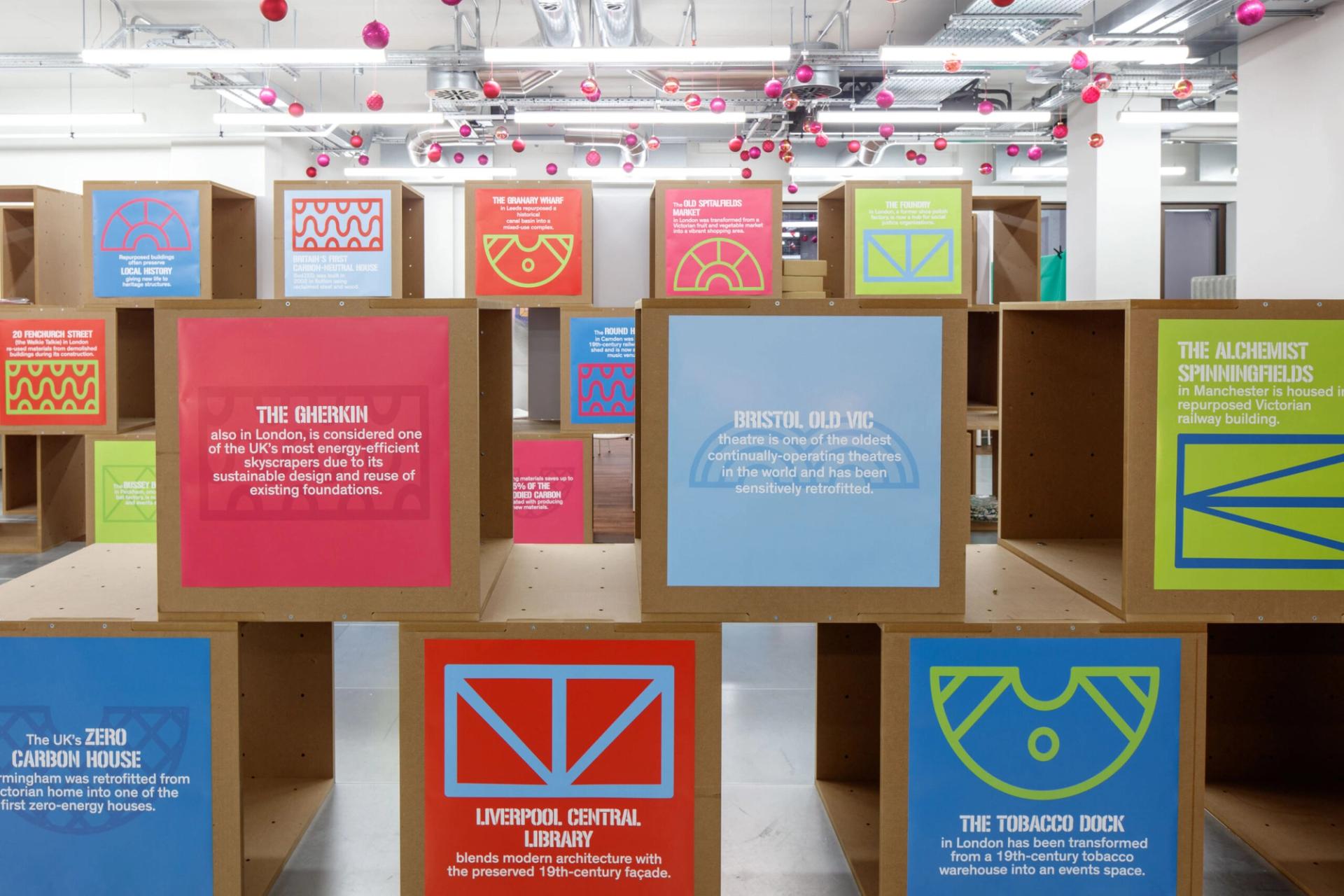 Colourful display boards with bold geometric graphics and text about London landmarks are mounted on modular spruce plywood cubes in a bright, modern exhibition space with pink ornaments hanging from the ceiling.