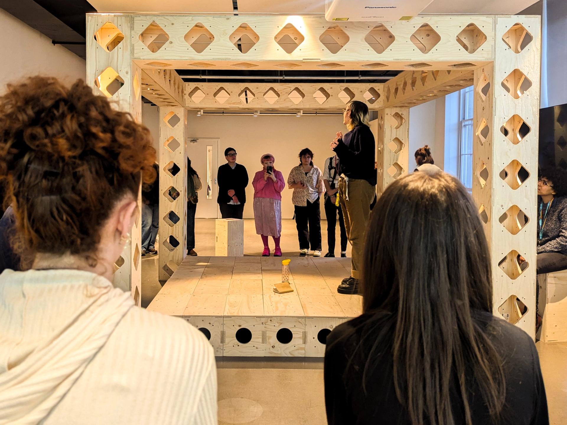 A group of people watch a woman speaking inside a large modular spruce art installation. Some sit in the foreground, while others stand around the structure, listening attentively. A yellow flower in a vase is placed on the floor inside.