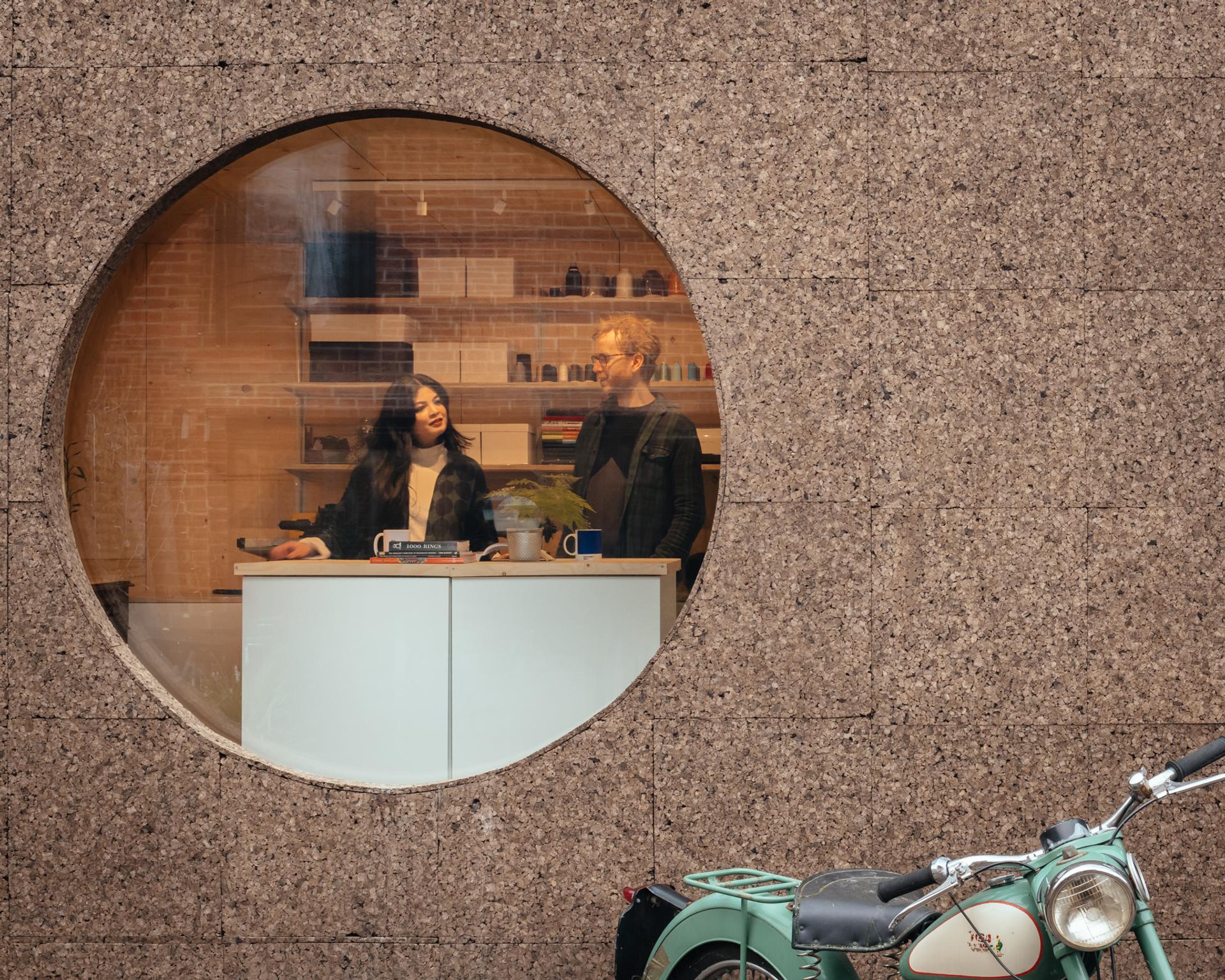 Two people stand behind a counter in a modern café, seen through a large round window set in a textured brown wall. A vintage green motorbike, reflecting the spirit of reuse and the circular economy, is parked outside in front of the window.