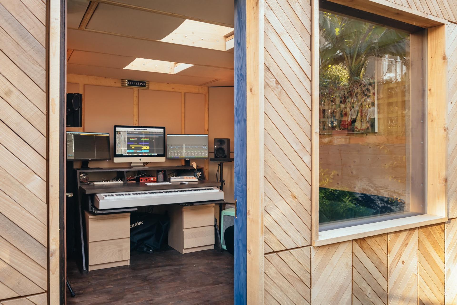A modern, sustainable wooden music studio with timber construction features a digital piano, multiple computer monitors, speakers, and audio equipment inside, viewed through an open door with sunlight streaming in.