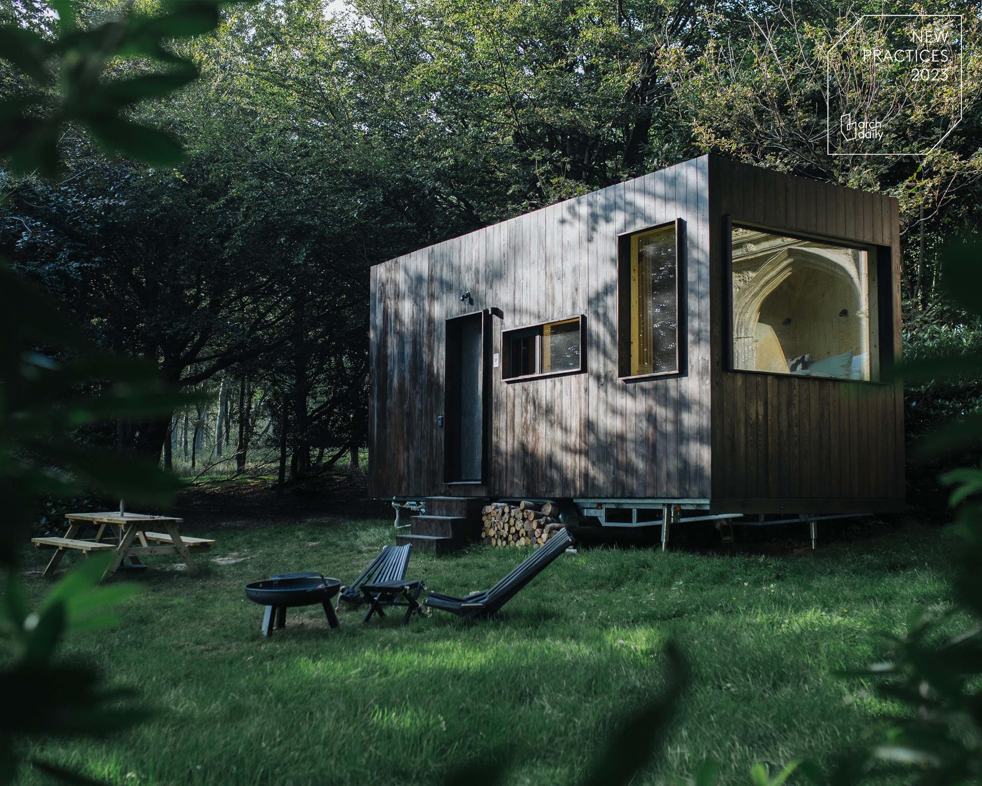 A modern spruce tiny house with large windows sits in a grassy clearing surrounded by trees. Nearby are outdoor chairs, a picnic table, and a fire pit, creating a peaceful, secluded outdoor retreat.