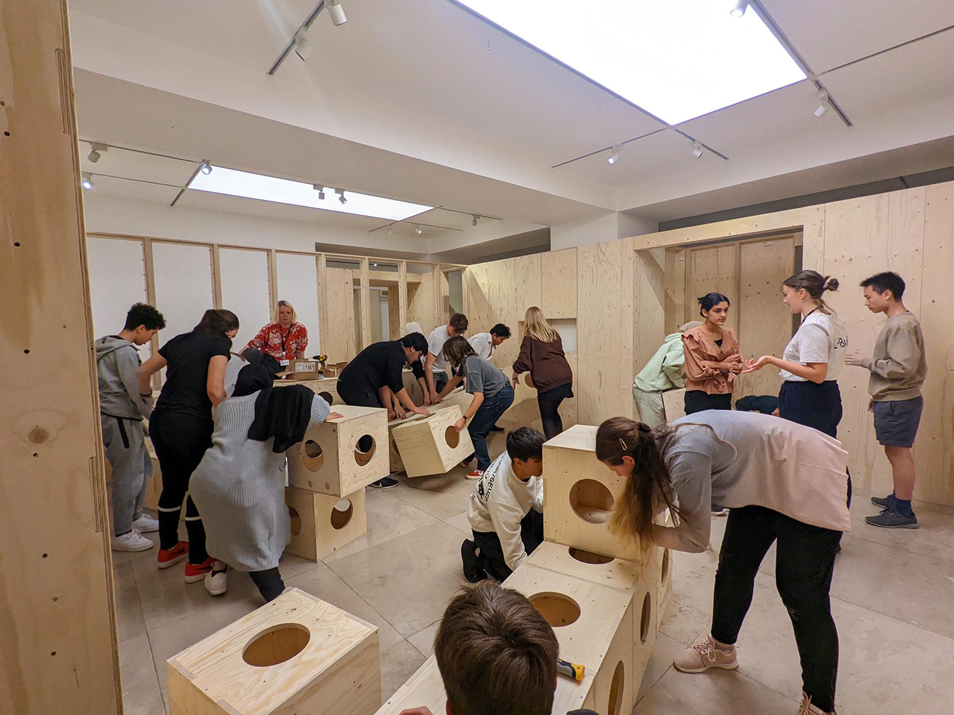 A group of people, including children and adults, assemble plywood boxes with circular holes in a brightly lit indoor space. Some work together whilst others observe or chat. The room features light wood walls and ceiling lights.