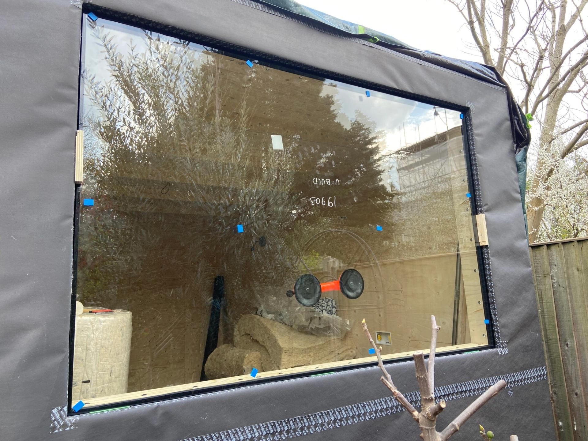 A large window with blue tape around its edges is installed in a partially constructed spruce wall. The reflection of trees shimmers in the glass, handwritten numbers mark the window, and construction materials are visible inside.