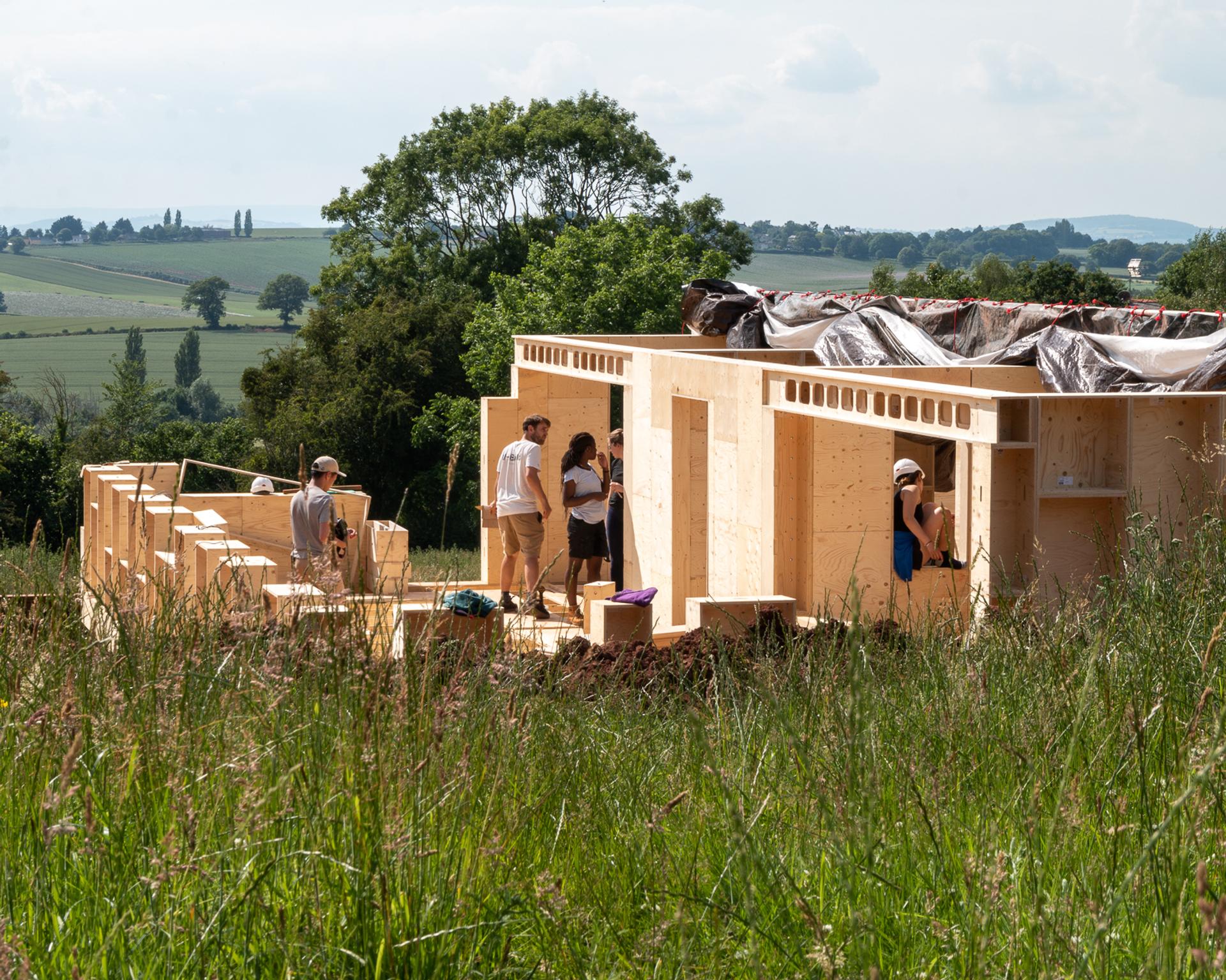 A group of people work together on building a wooden structure with sustainably sourced materials in a grassy field, surrounded by rolling hills and trees under a partly cloudy sky.