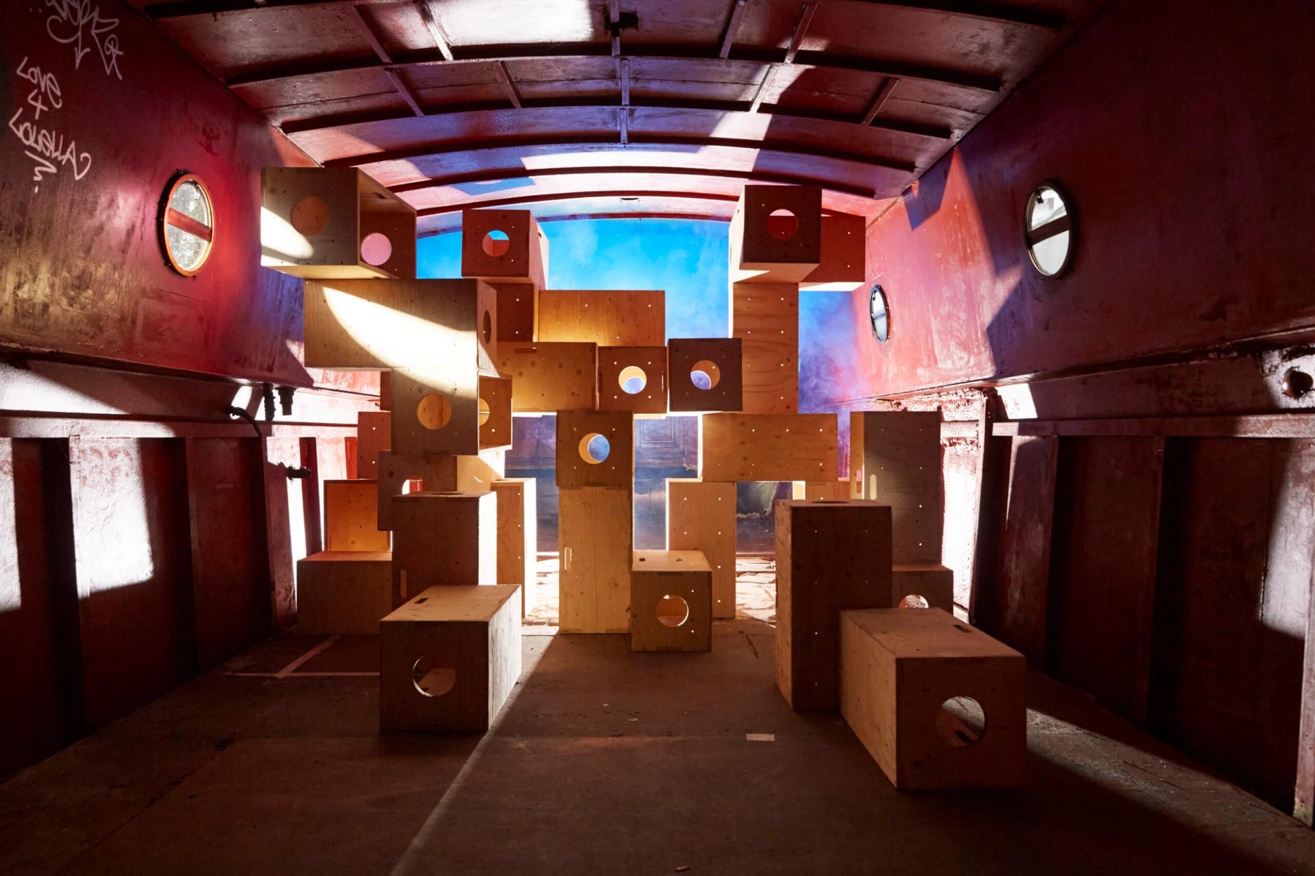 A cluster of modular plywood boxes of various sizes stacked in the center of a red-walled industrial space or cargo container. Bright, warm sunlight streams from the left, casting sharp shadows, while a cool blue light glows from behind the structure. Each wooden box features a circular cutout.