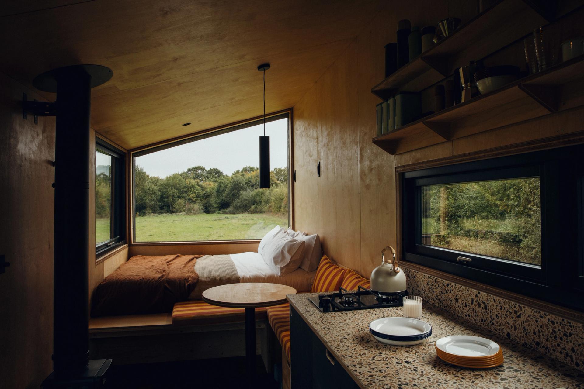 A cosy tiny house interior features spruce accents and a bed beside a large window overlooking green fields, a small table, and a kitchenette with a kettle on the hob. Open shelves hold crockery and cups as warm, natural light fills the space.