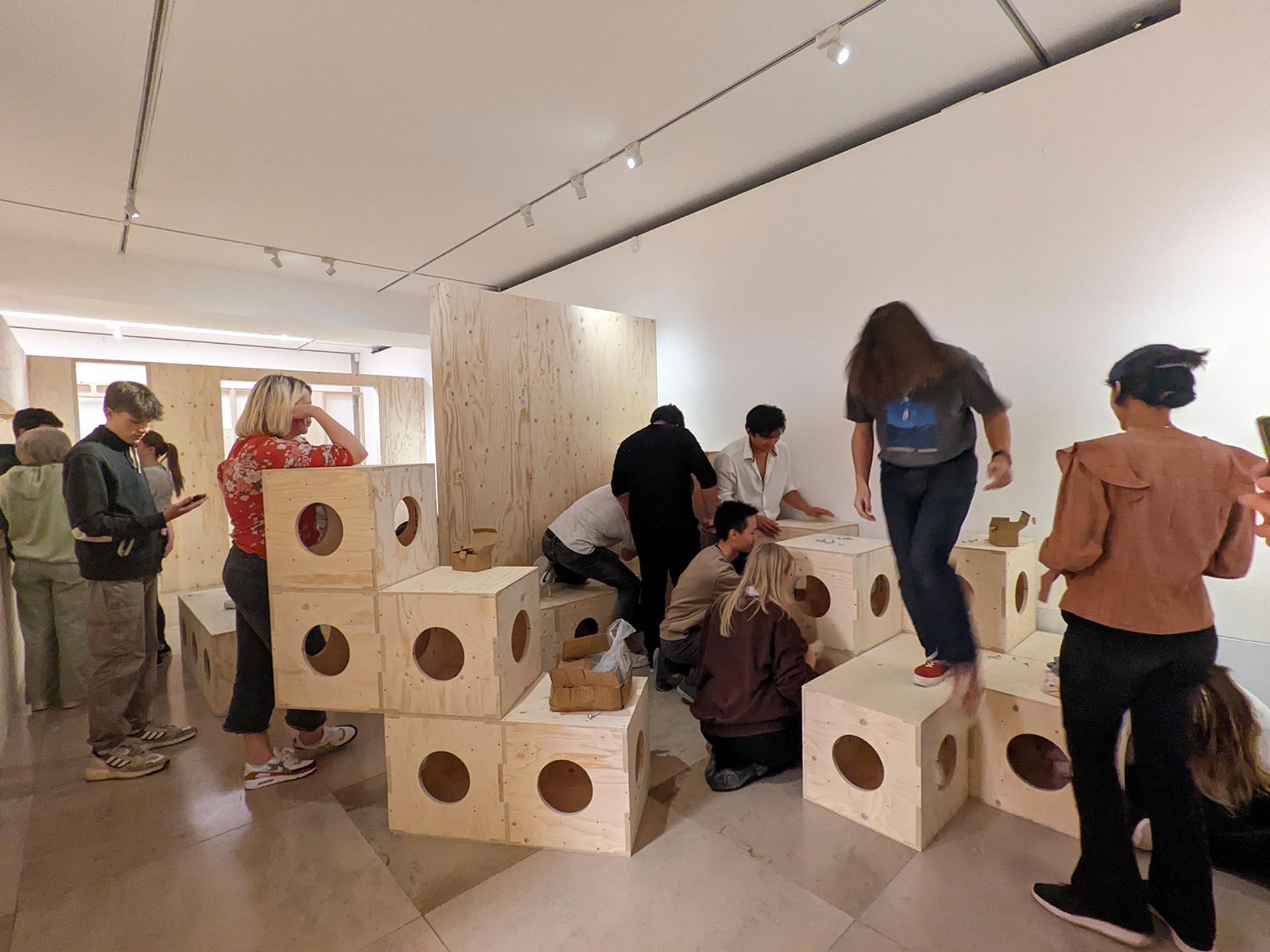 A group of people interact with large modular birch blocks featuring circular cut-outs in a bright, modern gallery space. Some sit or kneel on the sustainable blocks, while others stand or observe.