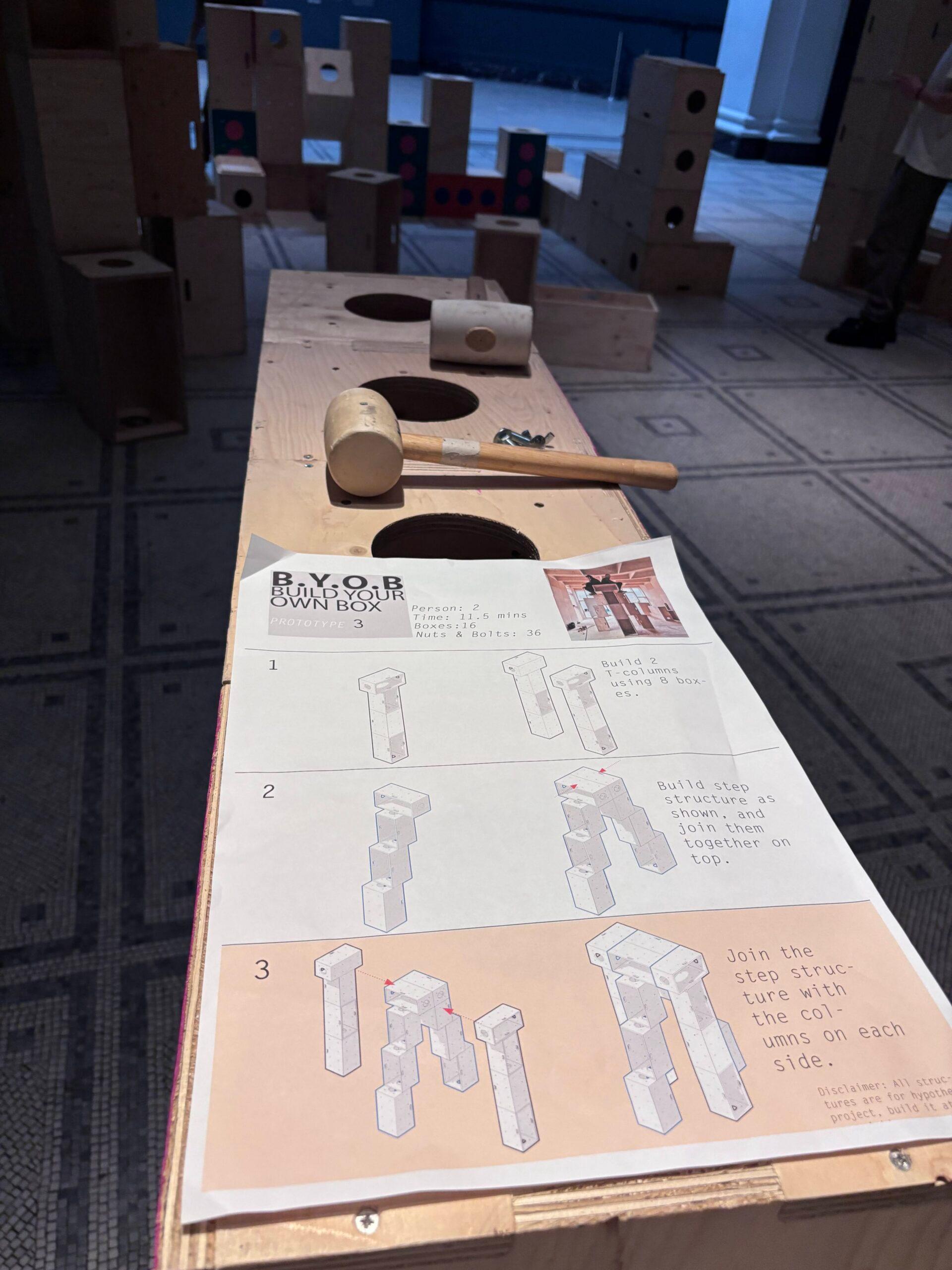 A wooden workbench with holes, a mallet, and printed step-by-step construction instructions—part of the Upstart initiative at the V&A—invites visitors to build structures for activism; wooden blocks and structures are visible in the background on a tiled floor.