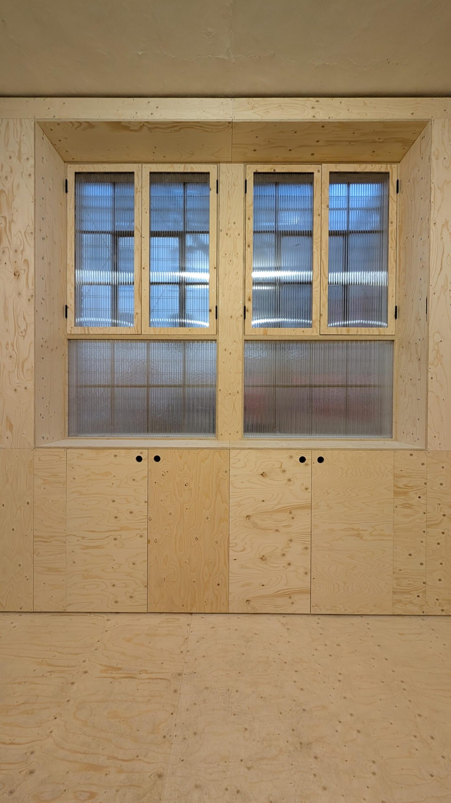 A wall with two rectangular windows above built-in cupboards, all made of light-coloured birch plywood. The ribbed, frosted glass and natural wood grain highlight the appeal of timber construction in this inviting space.