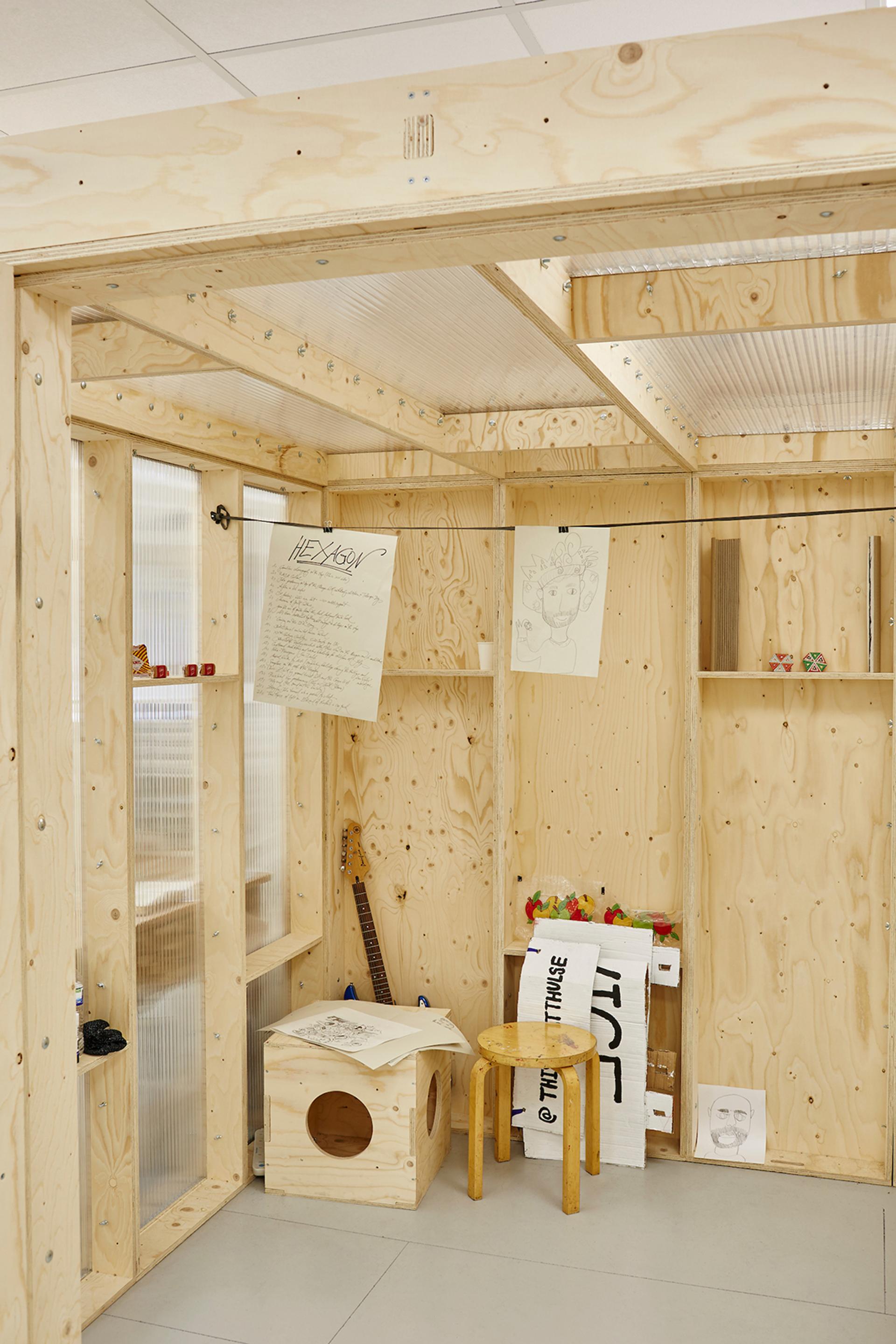 A small room with unfinished plywood walls and modular shelves holding scattered small items, hanging sketches, a wooden stool, a box with a hole, and various art supplies, giving the space a creative, workshop-like feel with hints of timber construction.