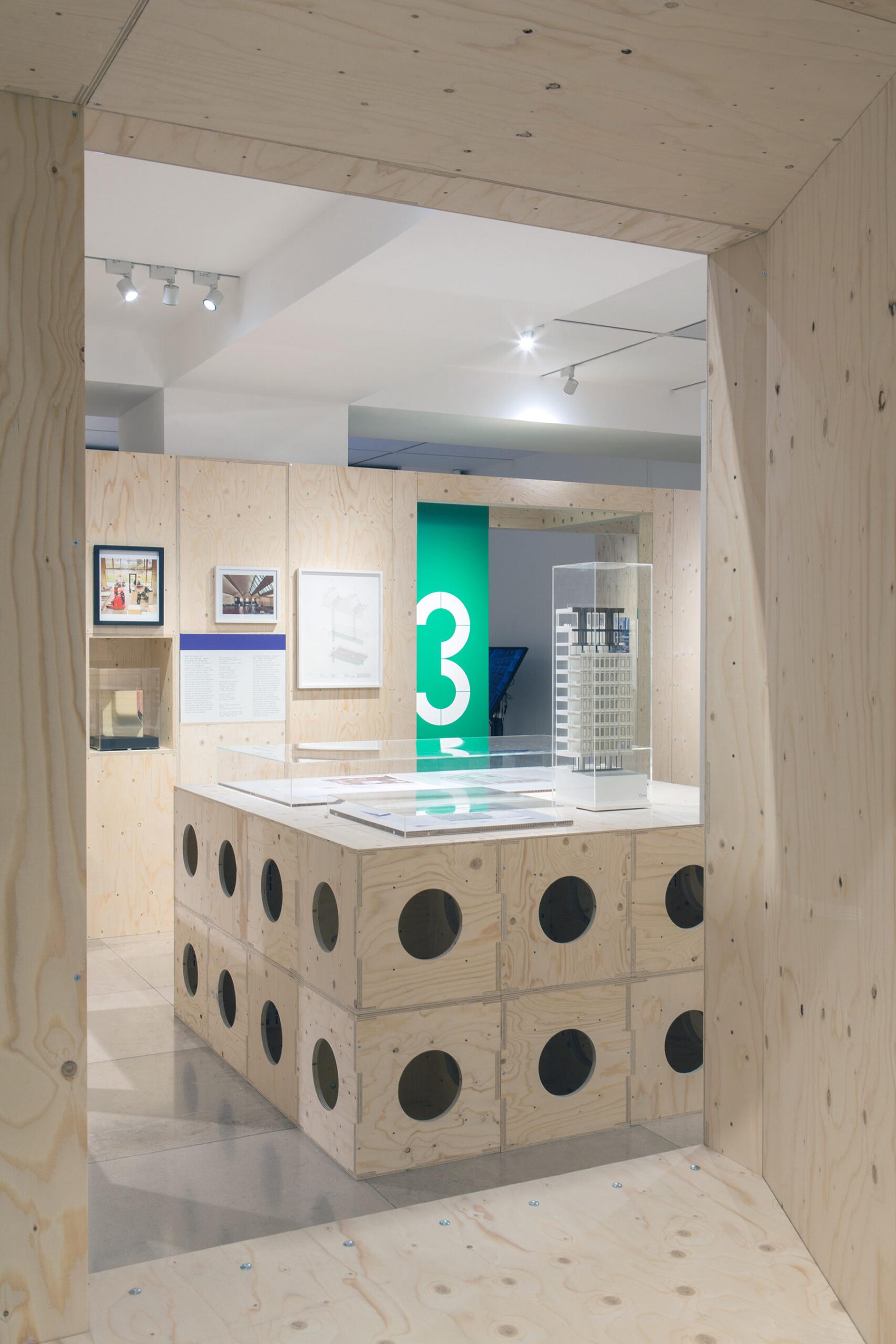 A modern exhibition space with light wood plywood walls and a display table featuring circular cut-outs. Architectural models and framed images are showcased, highlighting sustainable design, with a green sign labelled “3” in the background.