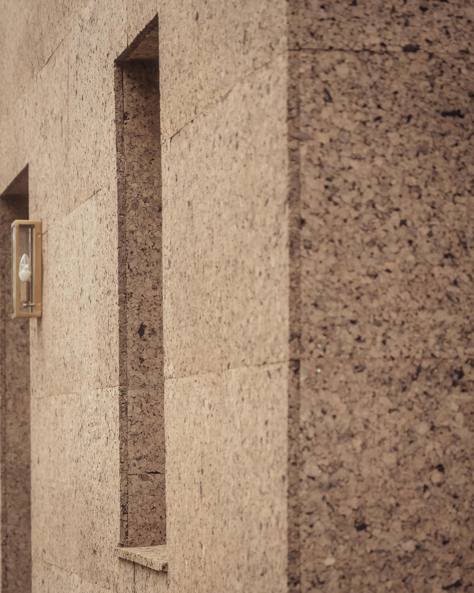 A wall covered in textured, tan cork cladding with a vertical recessed niche and a gold-toned wall light.