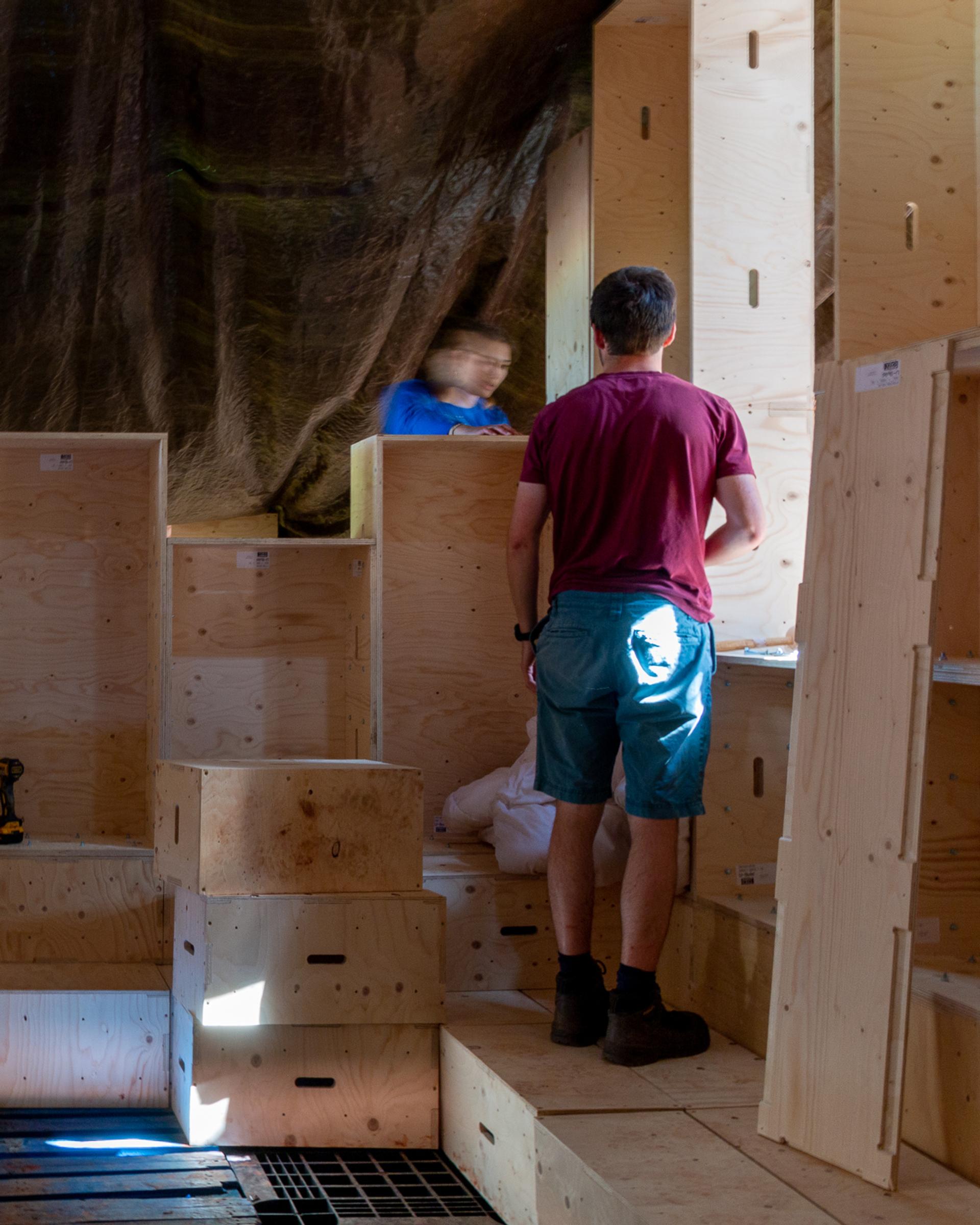 A man in a red shirt and blue shorts stands among large wooden crates, highlighting the potential to reuse materials. Another person is blurred in the background, with natural light streaming in from the right.