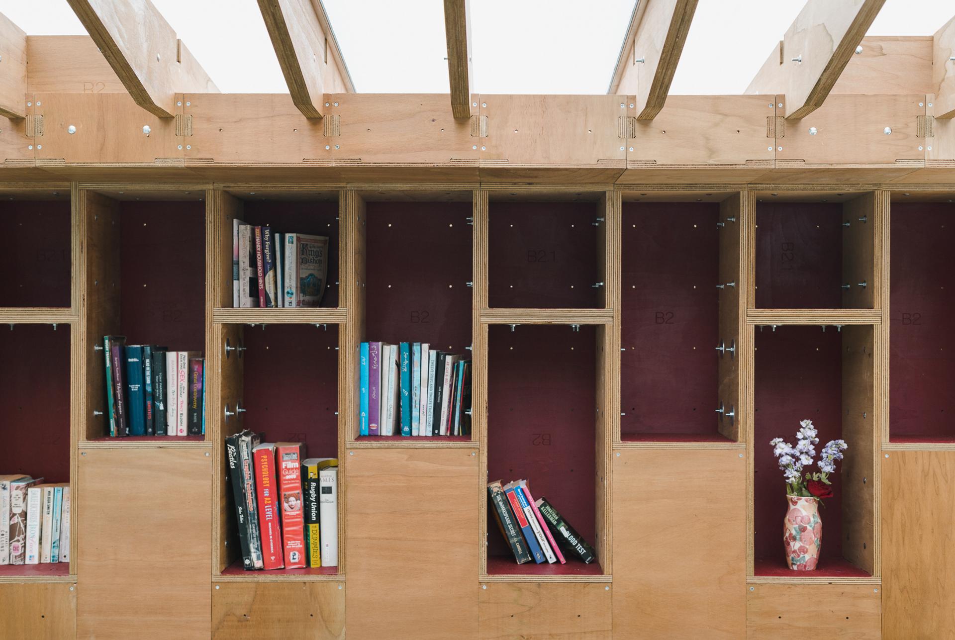 Wooden bookcases with open compartments, some holding books arranged vertically and horizontally. A pink floral vase with small purple flowers sits on the bottom right shelf. Exposed beams are visible above. Sustainably sourced timber adds charm.