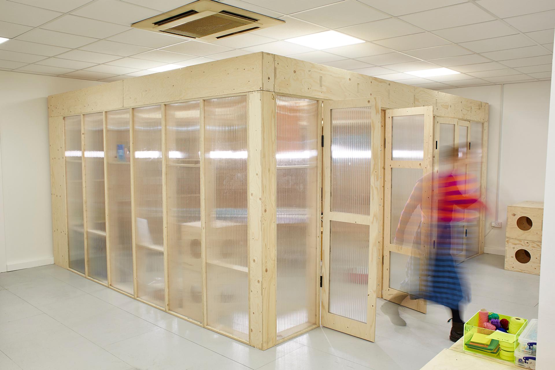 A person walks past a modern indoor structure featuring modular light wood and translucent panels, set in a bright room with white walls, a tiled ceiling, and a tidy, minimalist environment.