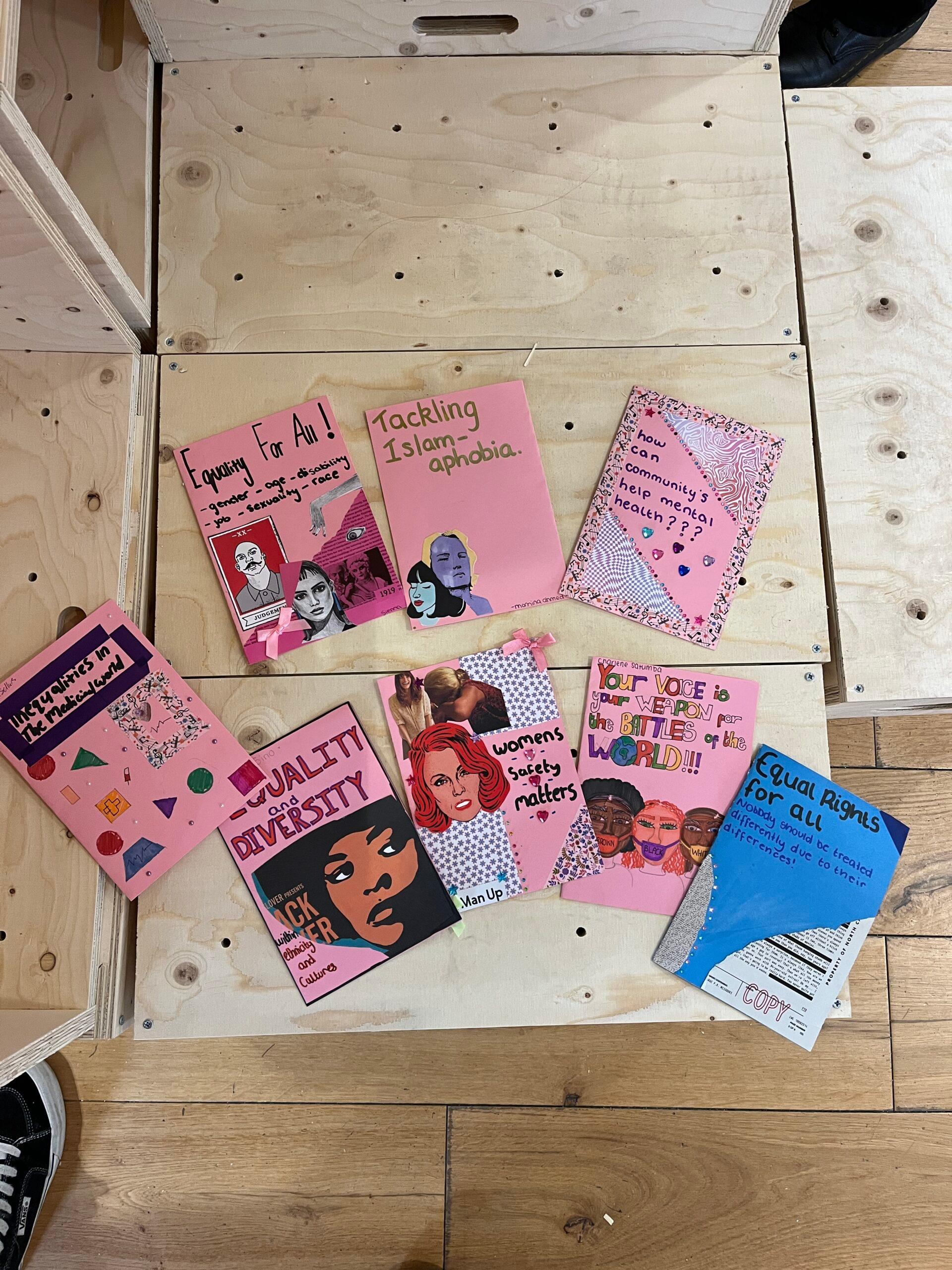 A collection of colourful zines on a plywood floor, covering topics like equality, diversity, Islamophobia, women’s rights, and community health, with hand-drawn illustrations and text on their covers.