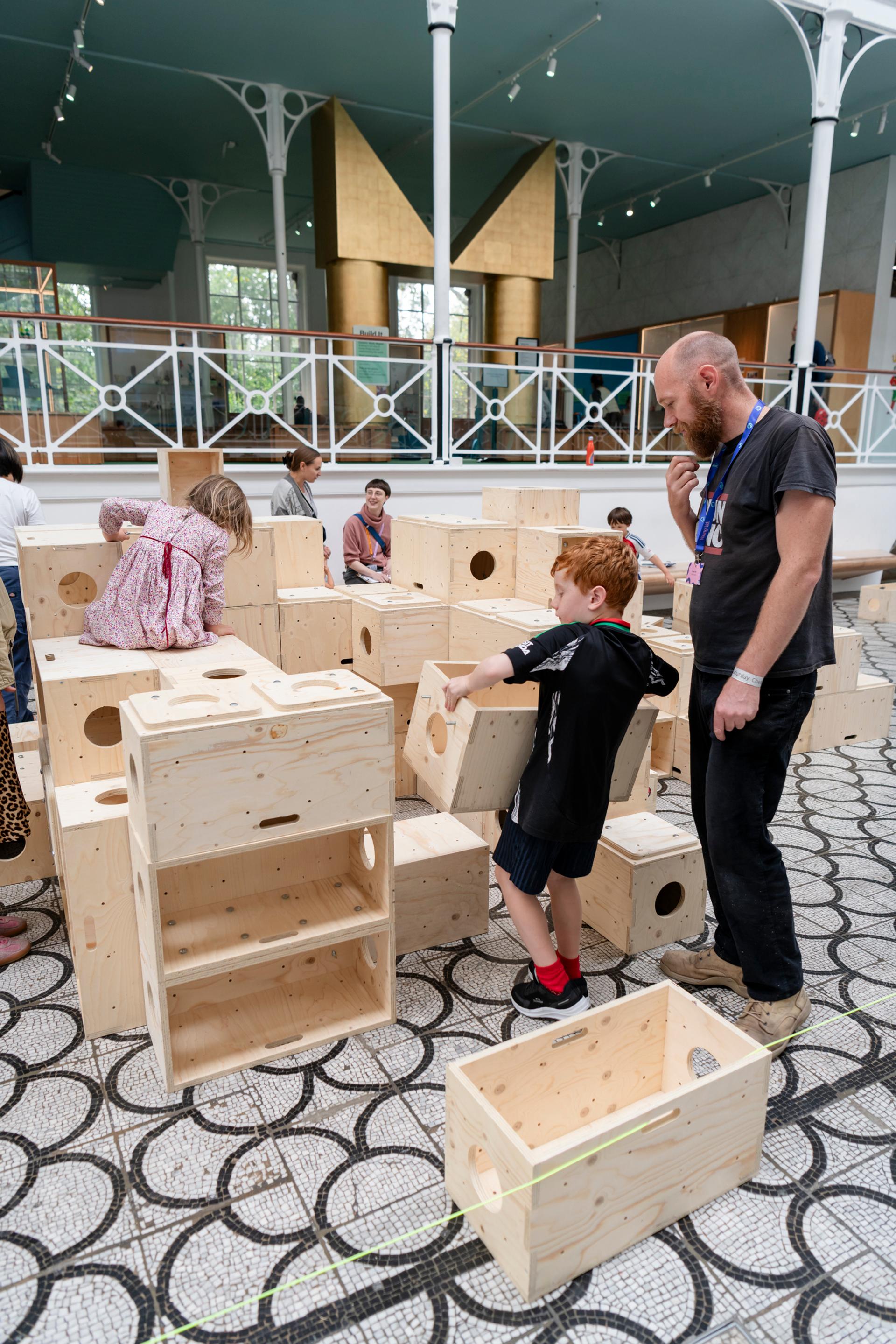Children and an adult interact with large U-Build wooden blocks in a spacious, indoor play area with patterned flooring and a modern, open structure in the background.