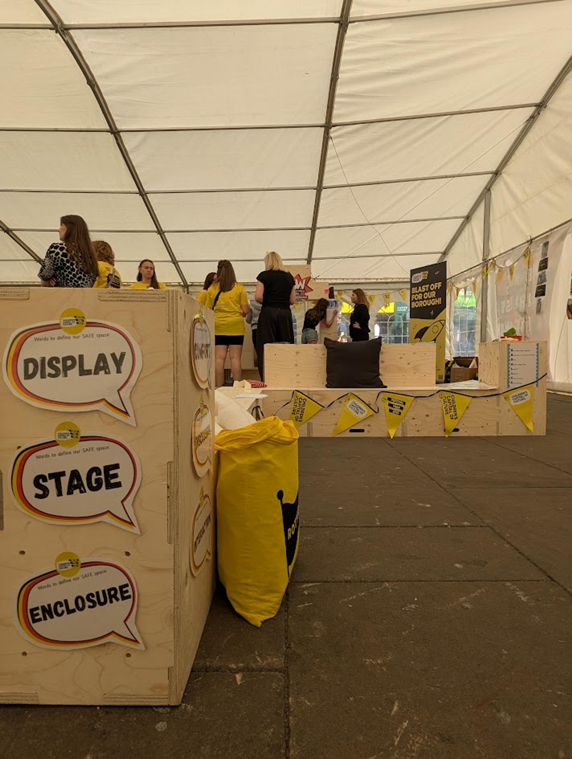 People stand and talk inside a large white event marquee. In the foreground, modular birch plywood crates labelled DISPLAY, STAGE, and ENCLOSURE sit among yellow bunting and bags. Informational signs are visible throughout the space.