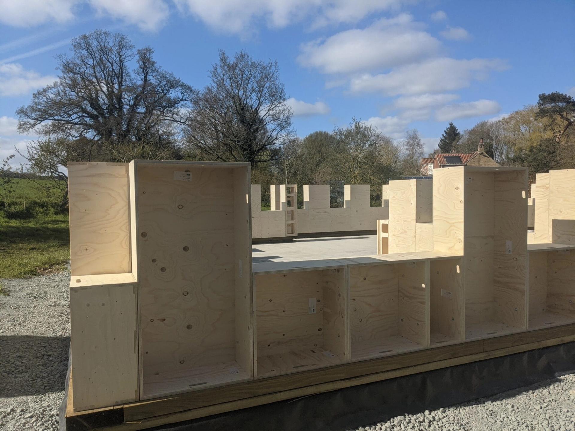 Partially constructed timber construction on a gravel foundation, featuring spruce beams, with trees and a house visible in the background under a partly cloudy sky.