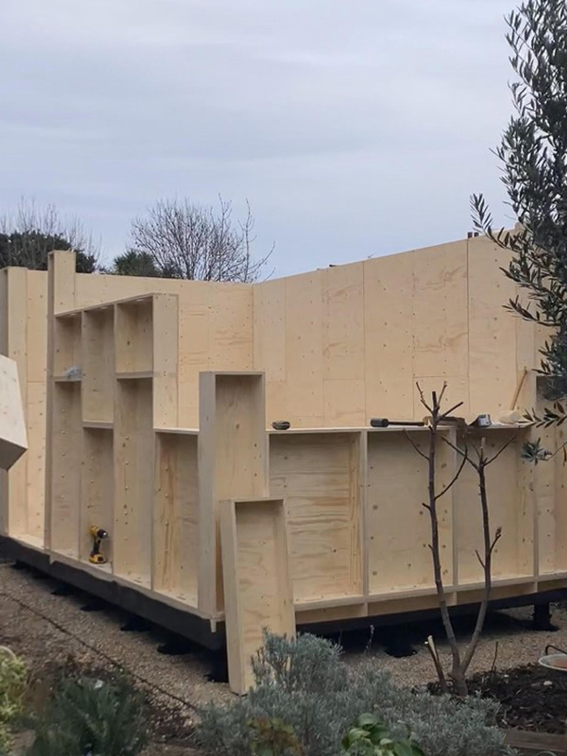 A partially constructed modular wooden building stands outdoors, with unfinished plywood walls and open sections. Construction tools rest on the flat surfaces, and bare trees and shrubs surround the structure under a cloudy sky.
