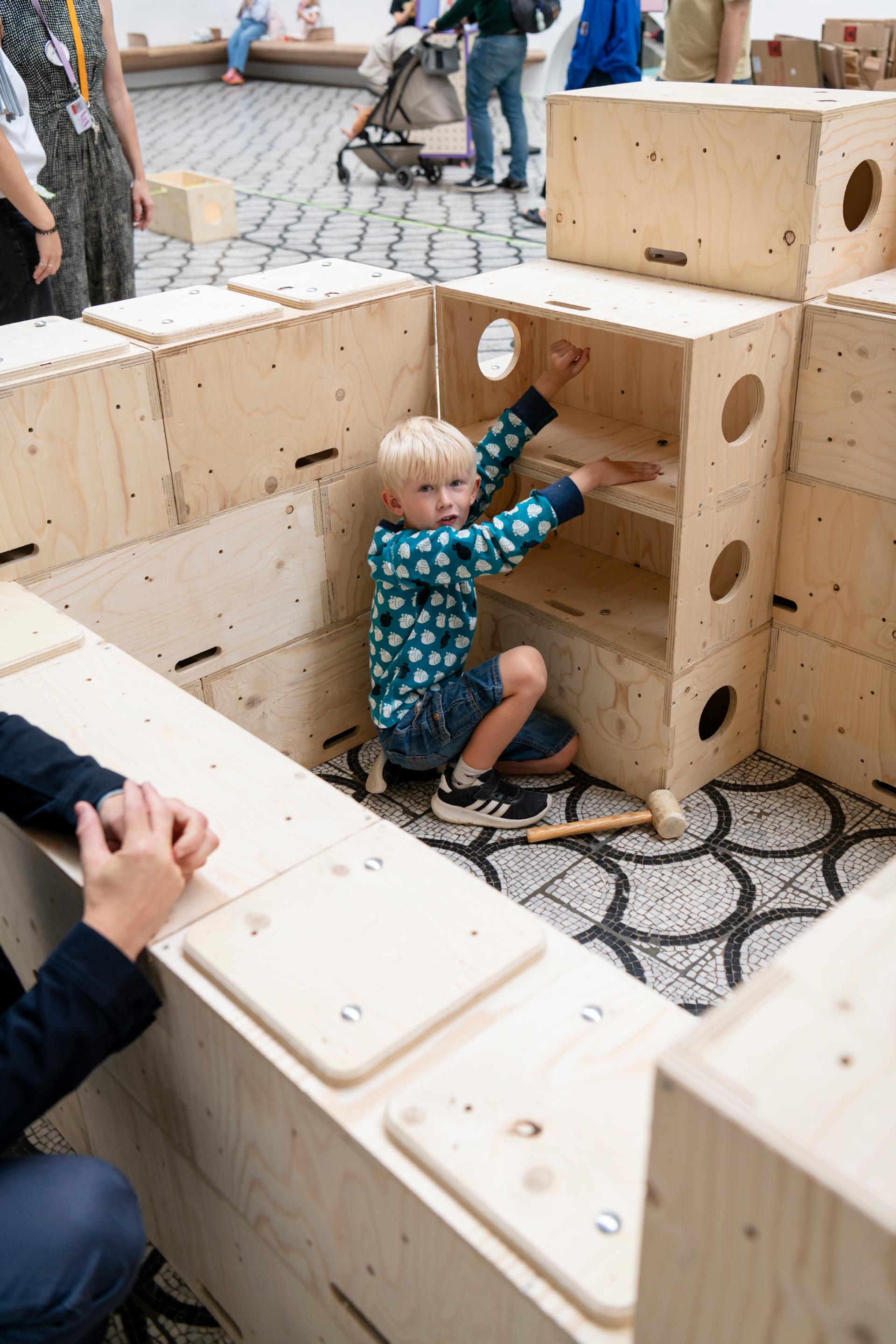 A young child with blond hair sits inside a U-Build structure made of large wooden blocks, looking up. Other people and children are around, and the floor is patterned with a black and white geometric design.
