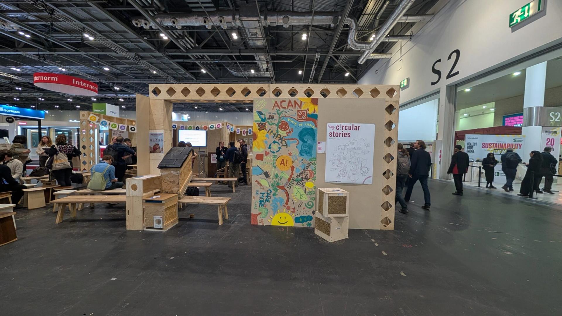 A colourful exhibition stand featuring a mural reading “ACAN” and “circular stories”, showcases sustainable timber construction with modular wooden structures, as people explore the spacious convention centre with high ceilings and exposed pipes.