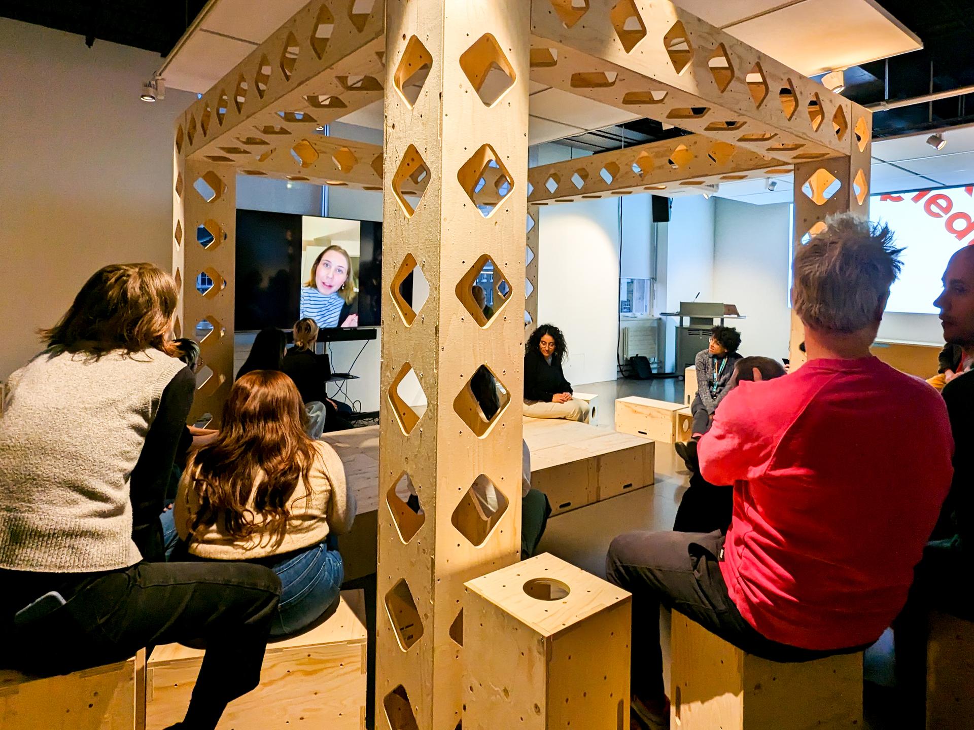 A group of people sit on wooden benches in a modern room featuring sustainable timber construction and geometric birch structures. They face a screen showing a video call with two people, suggesting a hybrid meeting or discussion.