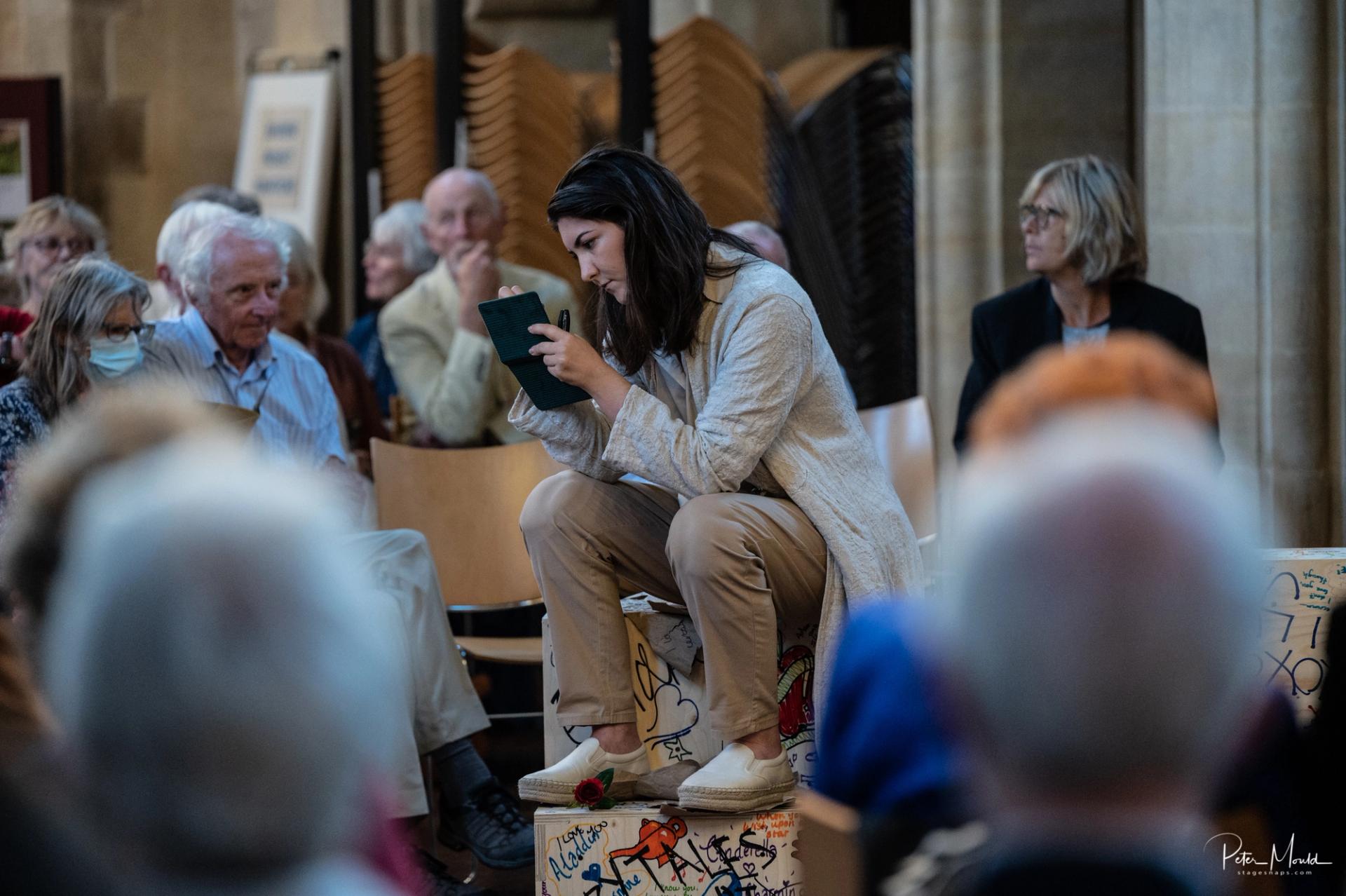A woman sits on a painted birch box, reading intently from a small book, surrounded by an audience in a hall. She is wearing a light-coloured outfit, and the atmosphere suggests a public event or performance.