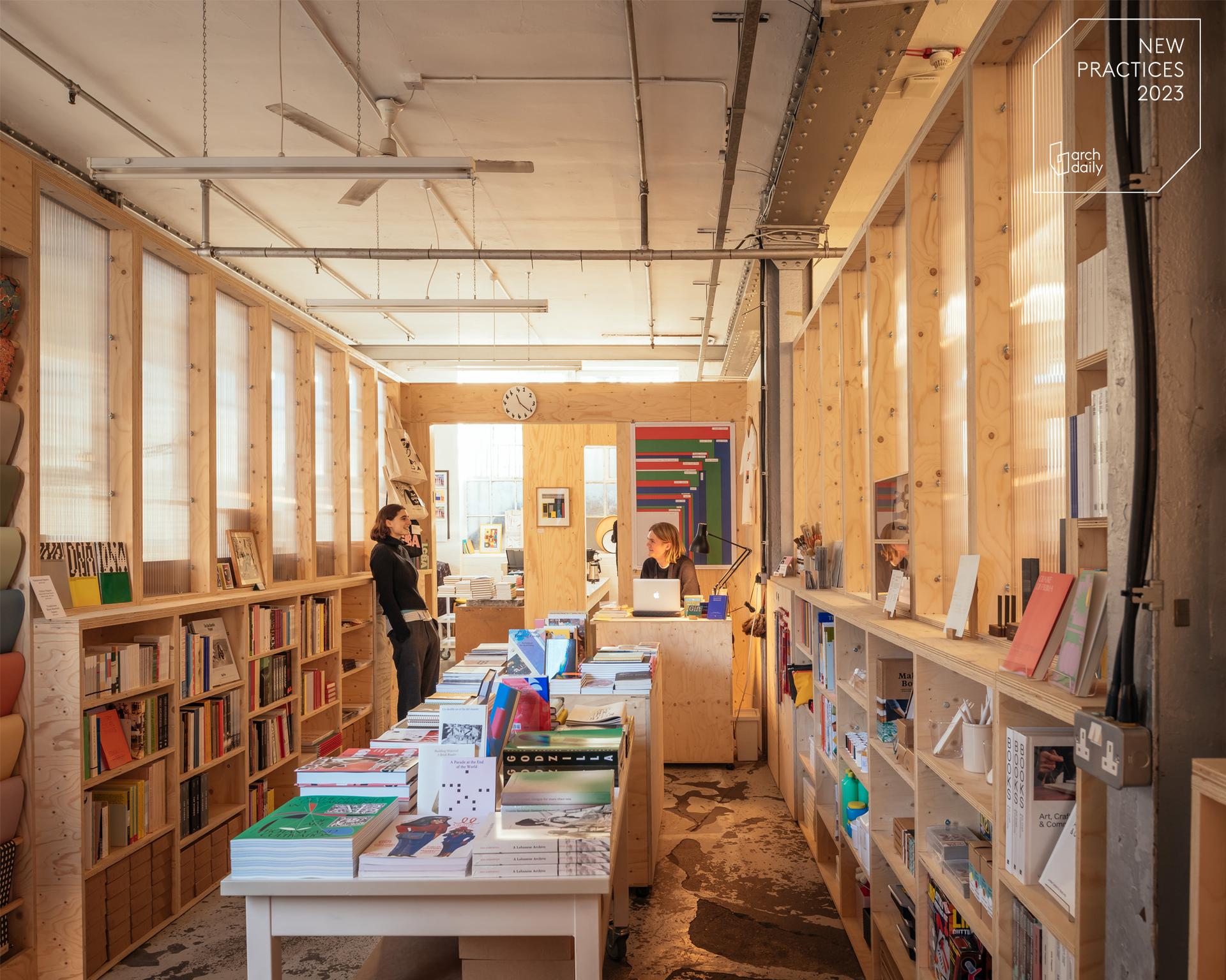 A modern bookshop with timber construction and wooden shelves filled with books, two people working at desks, and colourful books on display. Sunlight enters through large windows, creating a warm, inviting atmosphere. A clock and art hang on the back wall.