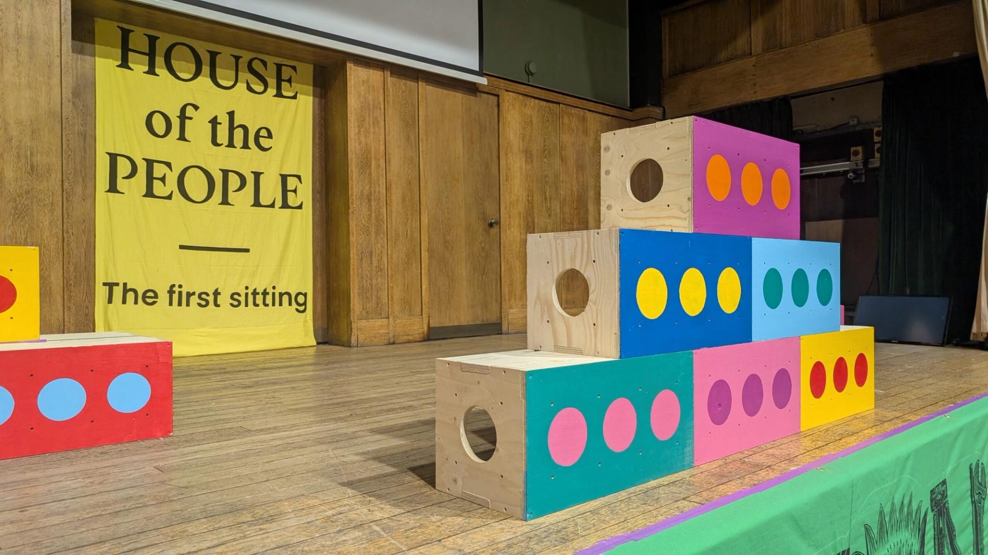 A stage with wooden walls displays a yellow banner reading HOUSE of the PEOPLE The first sitting. Colourful blocks with circles, made from sustainably sourced materials, are stacked at the front of the stage.