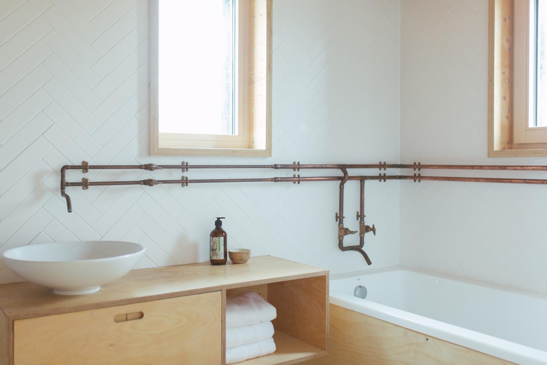 Minimalist bathroom with white herringbone tile, exposed copper pipes, a white vessel basin on a birch vanity, folded towels, and a built-in bath beneath two windows.
