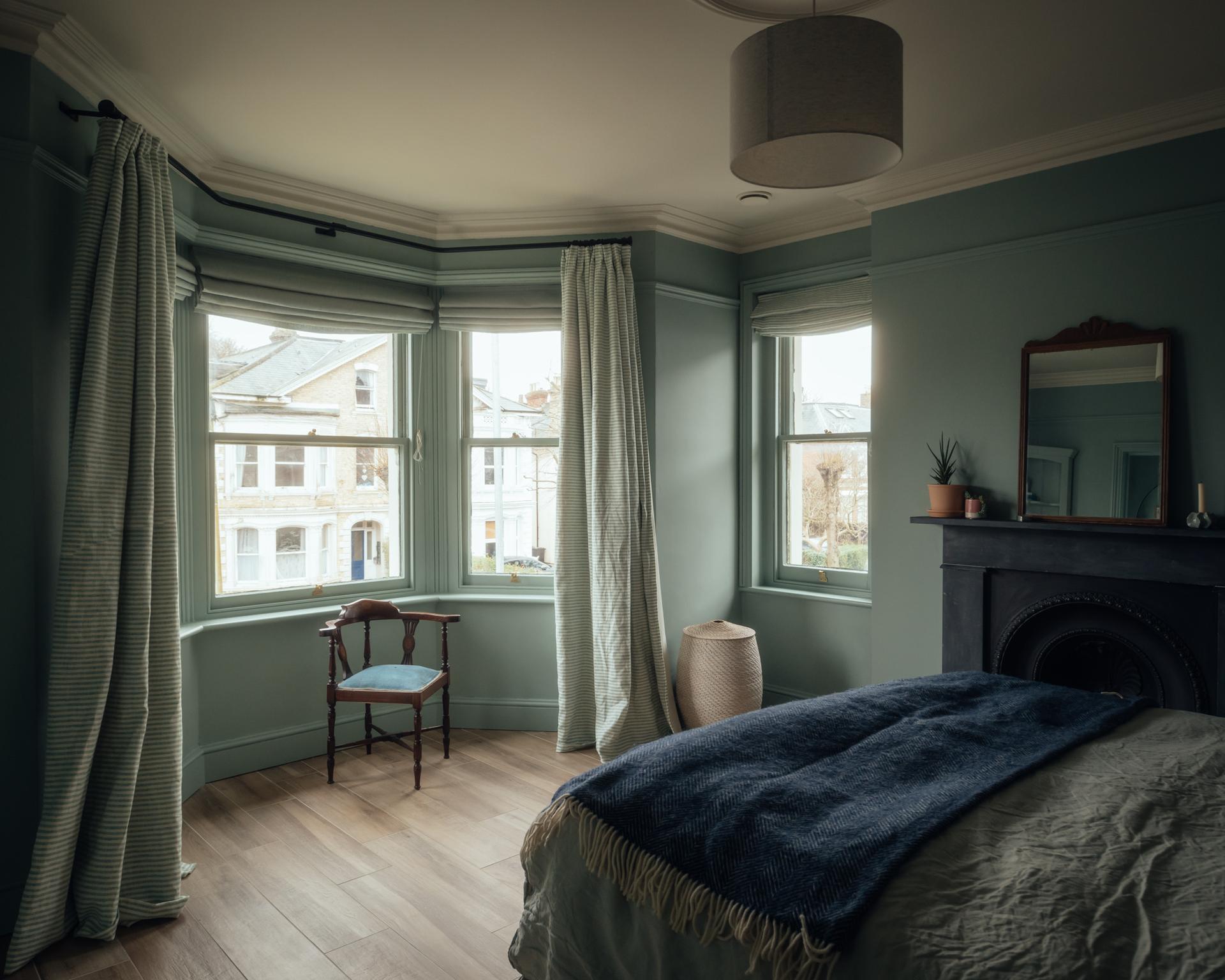 A bedroom with sage green walls, a bay window featuring a small wooden chair, and a bed with a dark blue throw blanket, all framed by timber architecture.