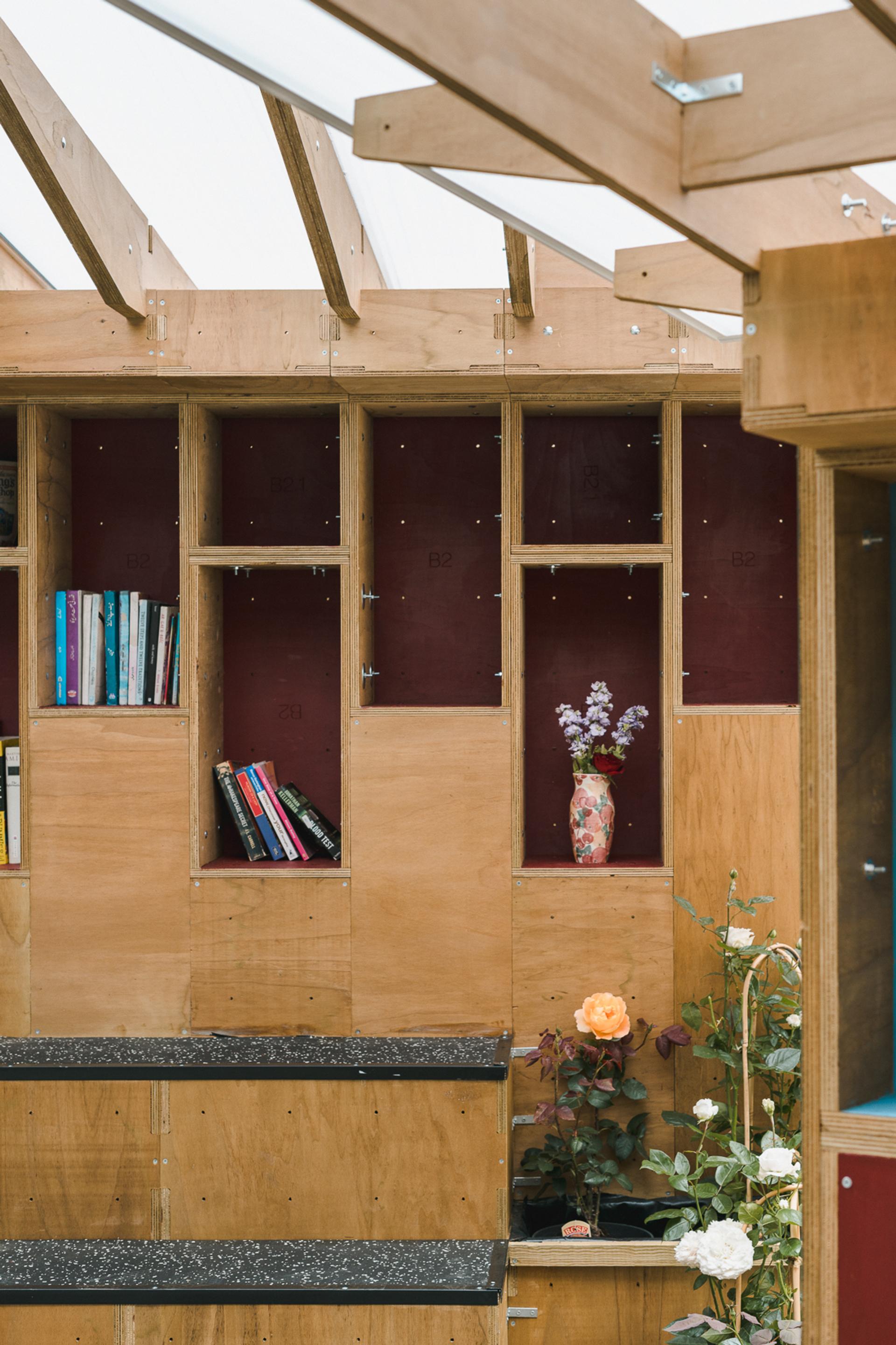 Wooden shelves with upcycled books and a vase of flowers rest against a maroon background. Below, various potted flowers and plants are arranged, bathed in natural light from a glass roof above.