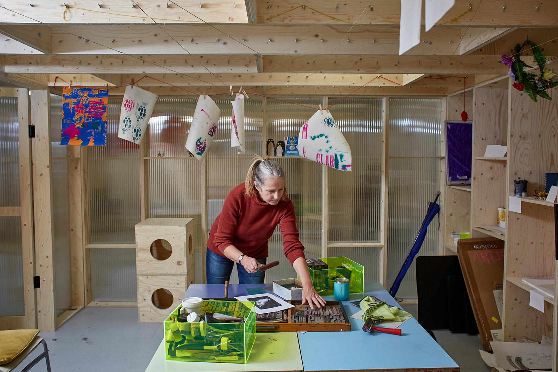 A woman in a red jumper works in a sustainable birch art studio, arranging prints and tools on a colourful table. Artworks and decorations hang above, with supplies organised in modular green bins around her.