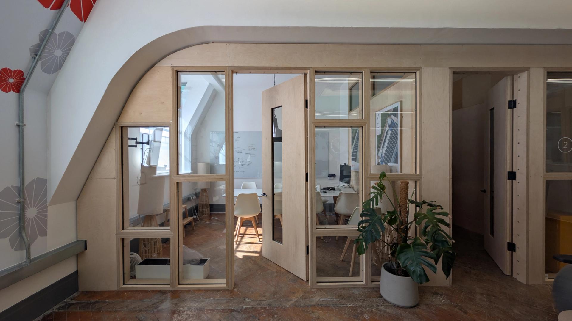 Modern office meeting room with large windows and a glass door, featuring white chairs around a table, a whiteboard, indoor plants, and natural light streaming in. Timber construction accents complement abstract art and flower patterns on the walls.