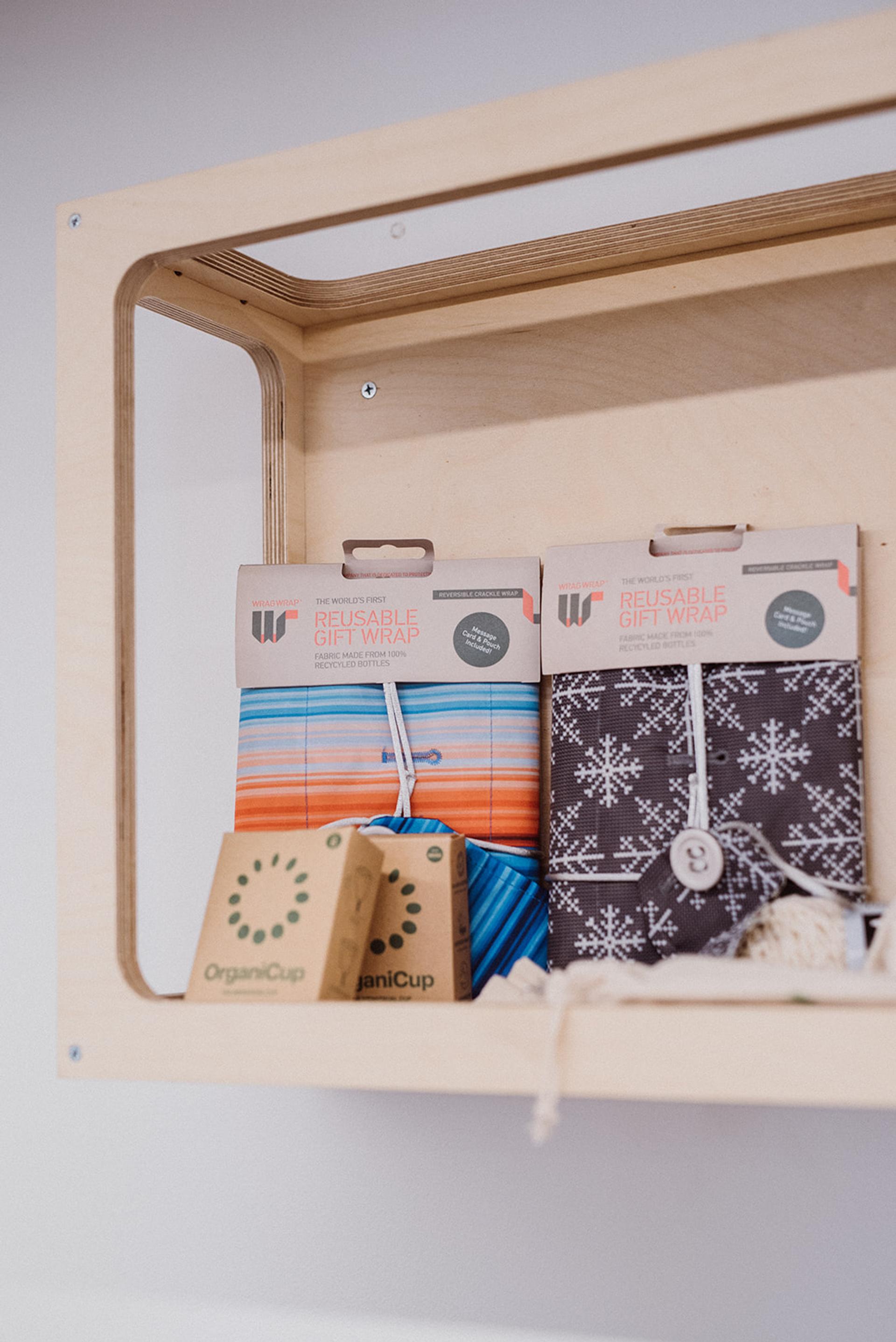 Two boxes of OrganiCup and two packs of reusable gift wrap—one with a blue and orange striped pattern, the other with a white snowflake design—are displayed on a birch plywood shelf.