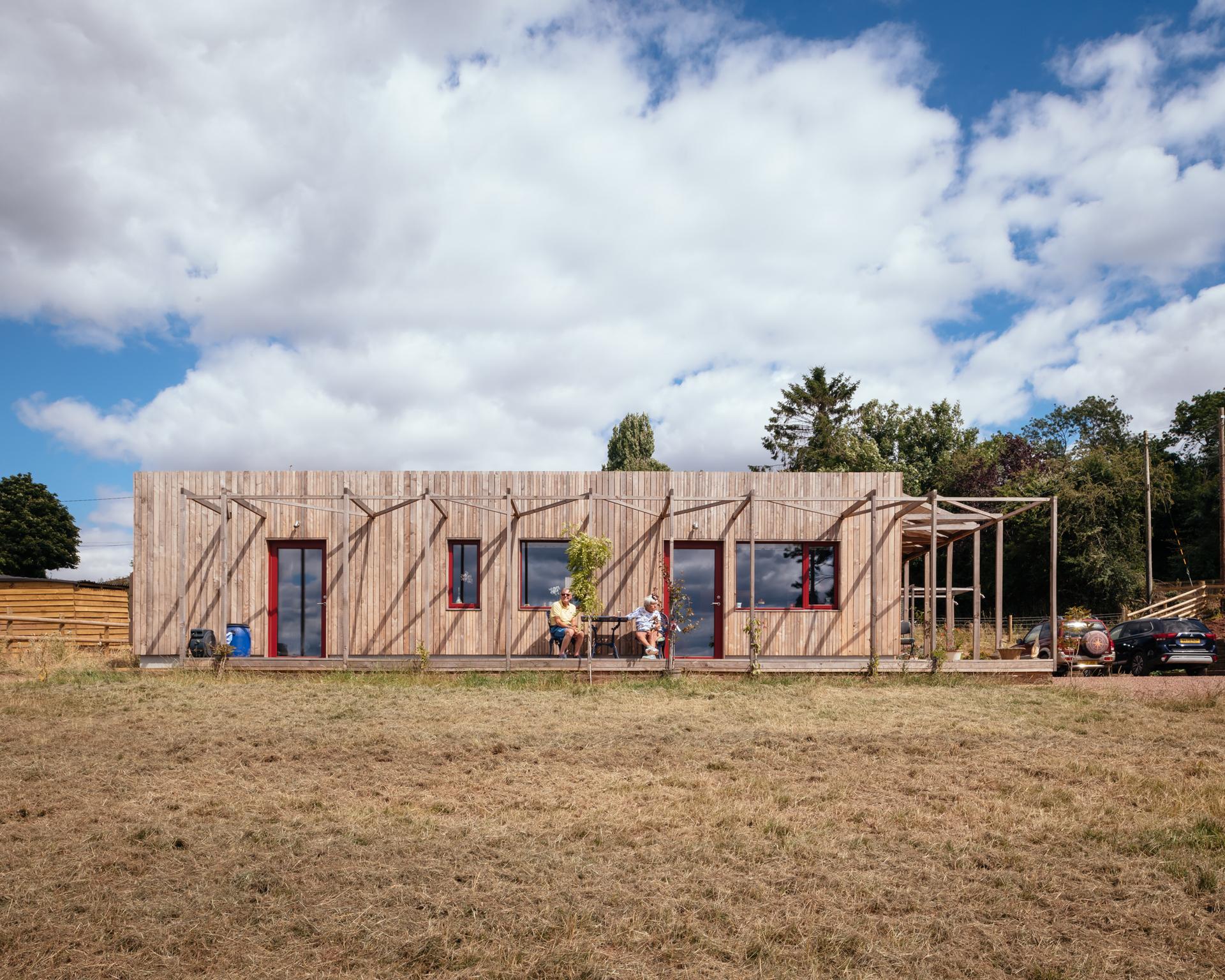 A modern modular wooden house with large windows sits under a partly cloudy sky; three people relax at a table on the front decking, and a car is parked to the right on dry grass. Trees and fencing are in the background.