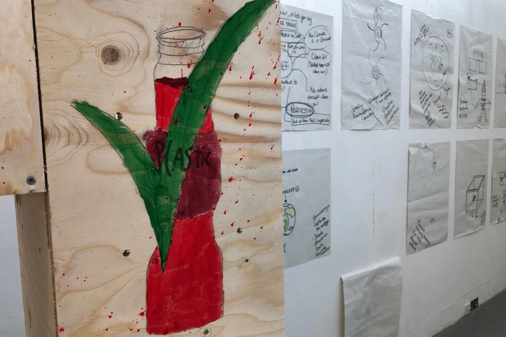 A wooden panel with a painted red bottle marked “PLASTIC” and a large green tick promotes the circular economy; sketches and diagrams about reuse are taped to a white wall in the background.