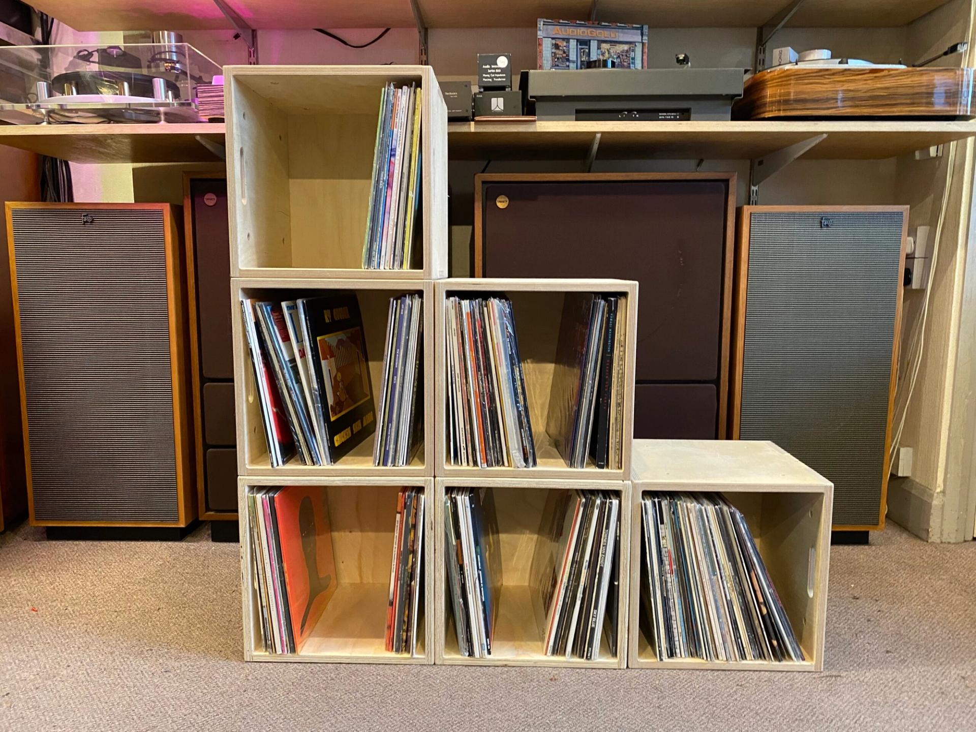 Six wooden crates, made from recycled materials, are filled with vinyl records and stacked on a carpeted floor. Behind them are two large speakers and shelves holding a record player and other audio equipment, promoting sustainability in music storage.