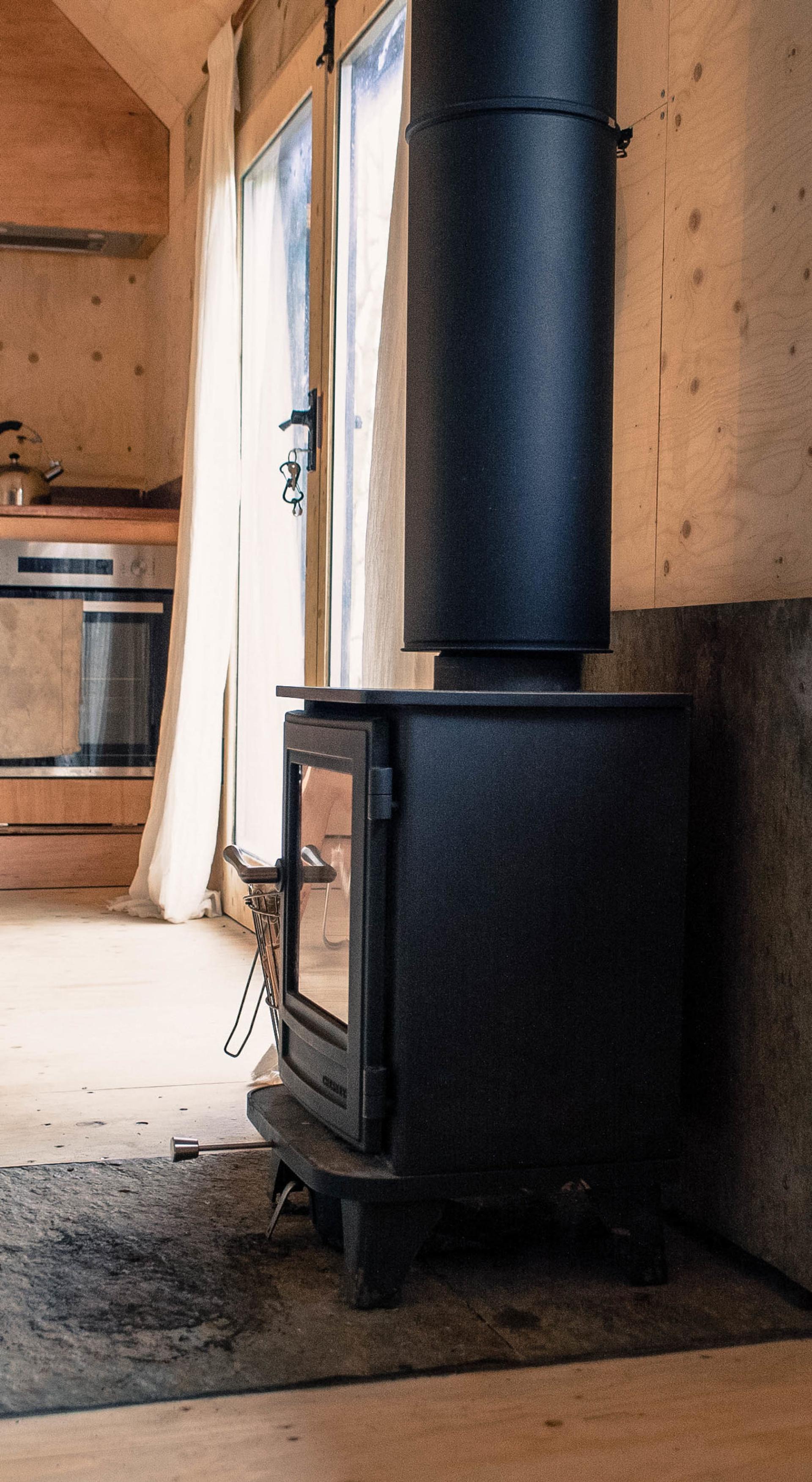A black wood-burning stove with a tall chimney stands on a stone hearth in a cosy wooden cabin, highlighting upcycle design with sheer curtains and a modern kitchen visible in the background.