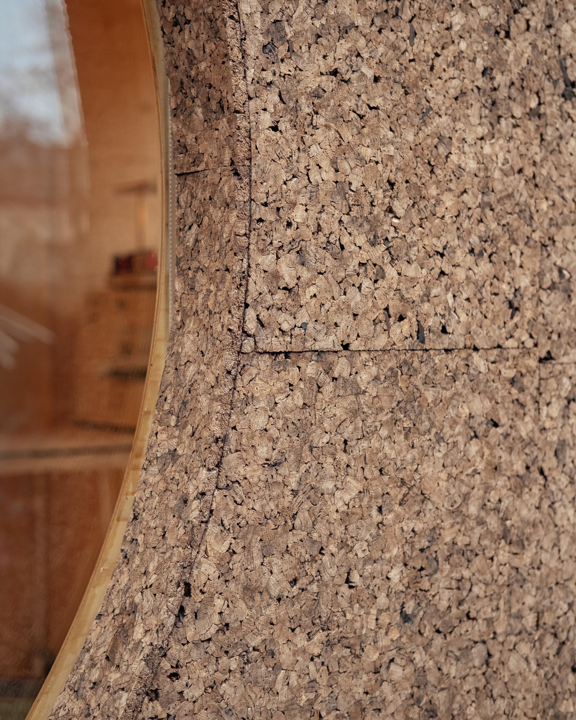 Close-up of a wall made from interlocking cork panels, featuring a round window with a wooden frame. The sustainable cork texture is rough and natural, whilst the window reveals a warm interior in the background.